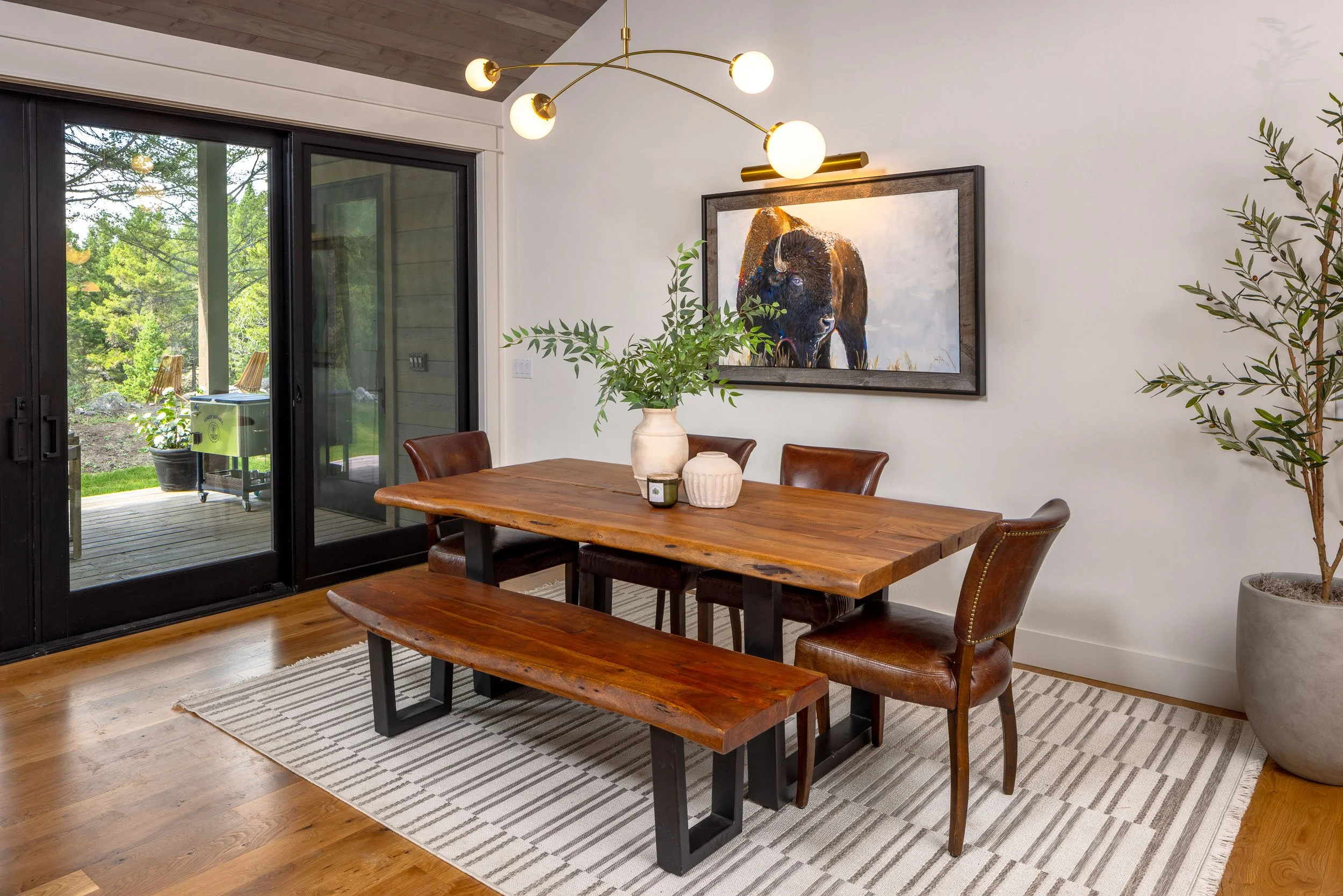 A dining room with a rustic wooden table, leather chairs, a bench, a large abstract painting of a bison, a modern gold chandelier, a sliding glass door leading to a deck with outdoor chairs and greenery, a striped area rug, and potted plants.