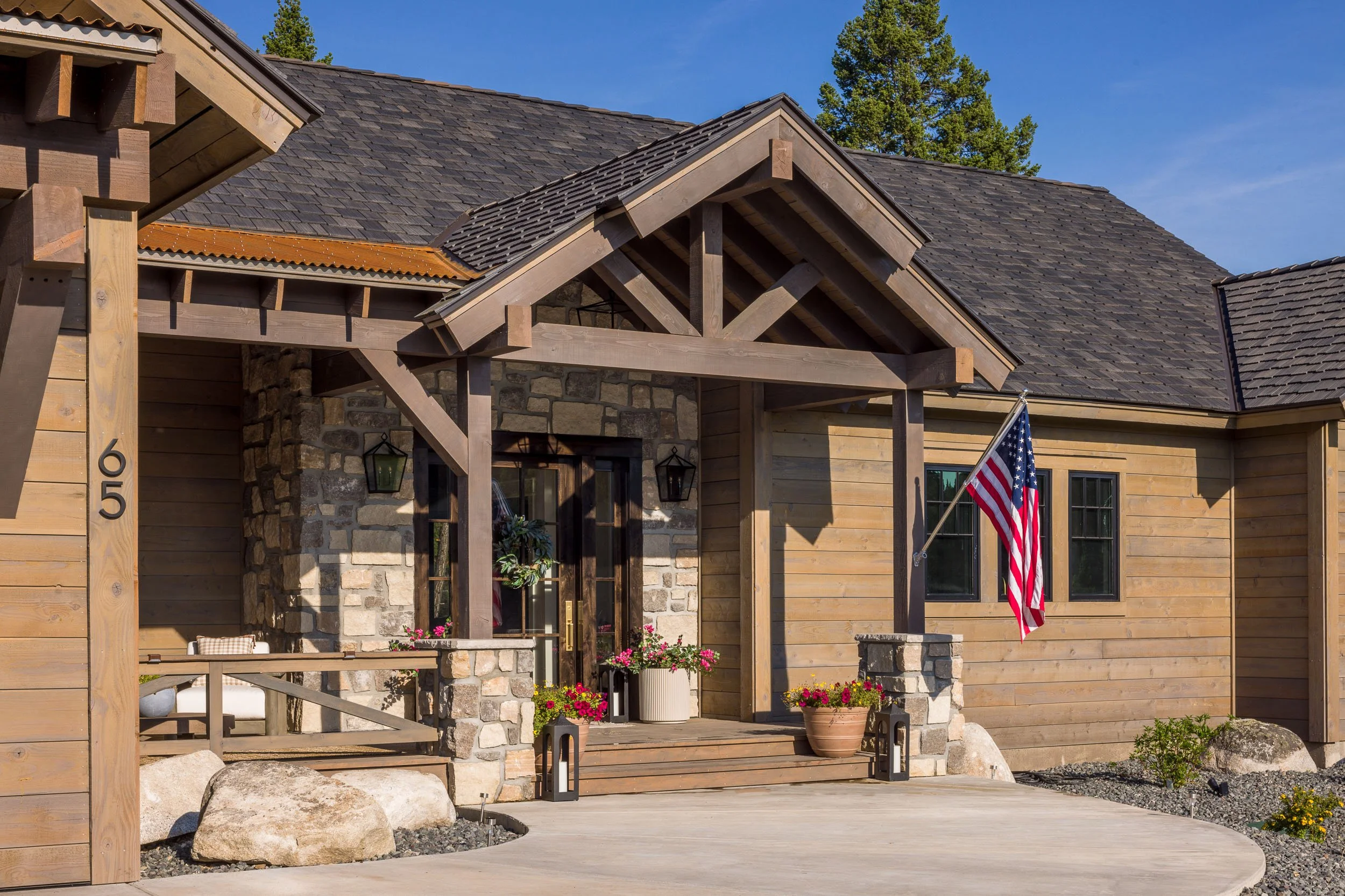 Front entrance of a house with wooden and stone exterior, American flag, potted plants, and lanterns, under a clear blue sky. Custom Home built in Montana by Edgell Building.