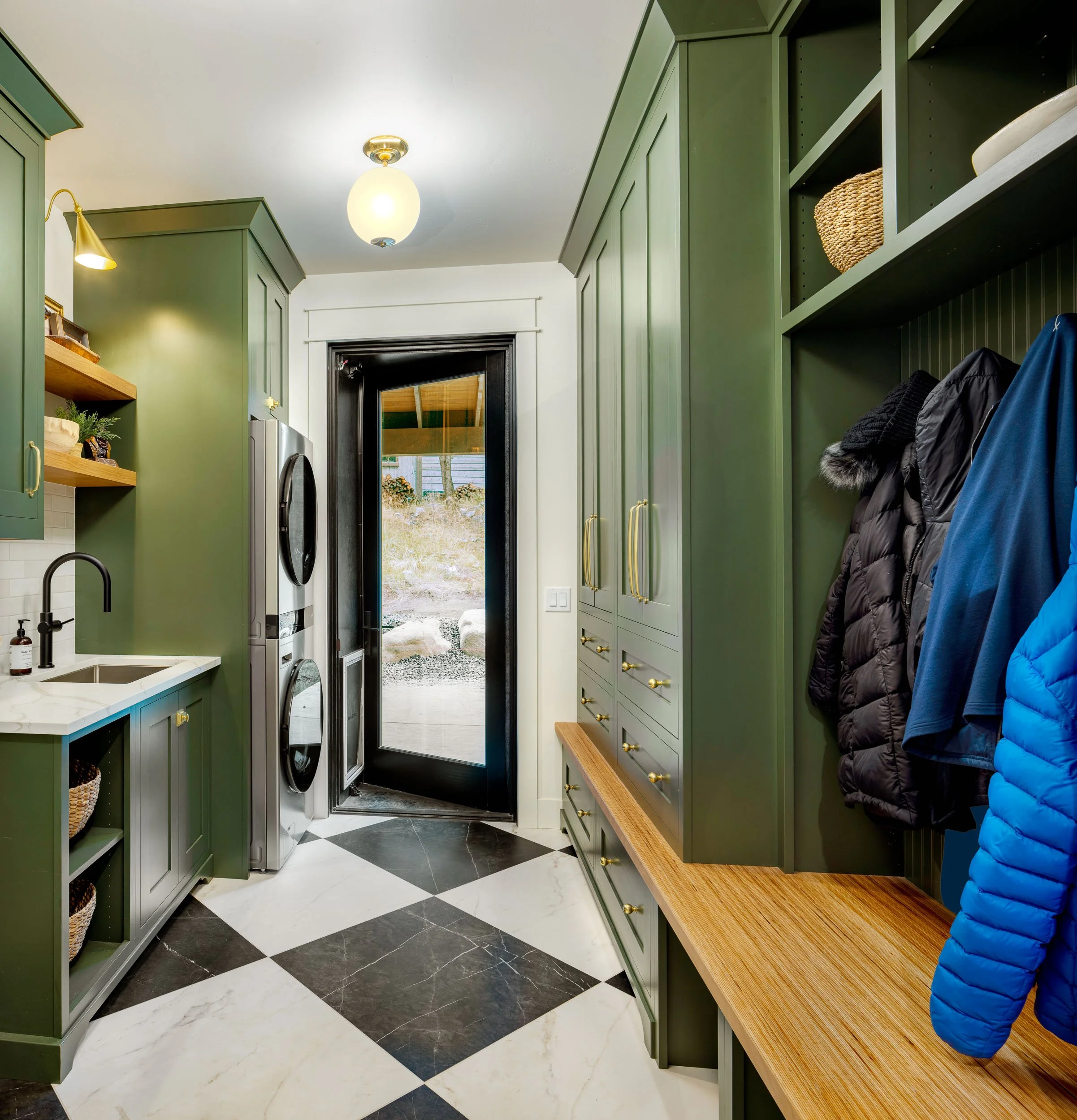 A laundry room with green cabinets, a washer and dryer stacked, black and white checkered marble floor, coat hooks with jackets, a door with a view outside, and wooden shelves. Custom Home built in Montana by Edgell Building.