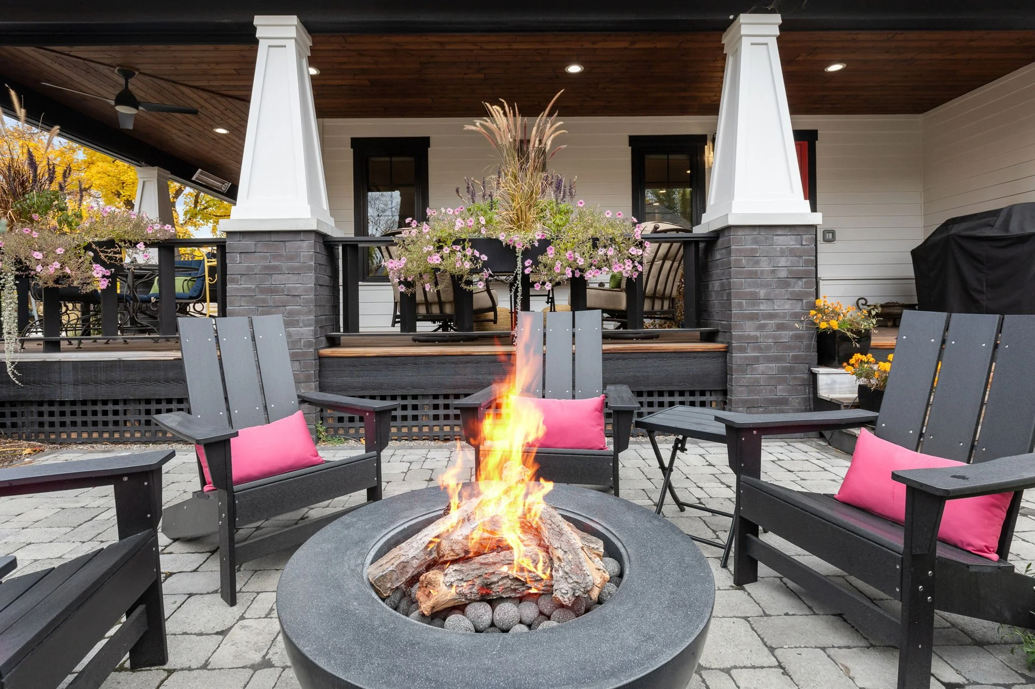 Outdoor patio with a fire pit surrounded by black Adirondack chairs with pink cushions, set in front of a house with a porch decorated with flower pots, plants, and patio furniture. Custom Home built in Montana by Edgell Building.