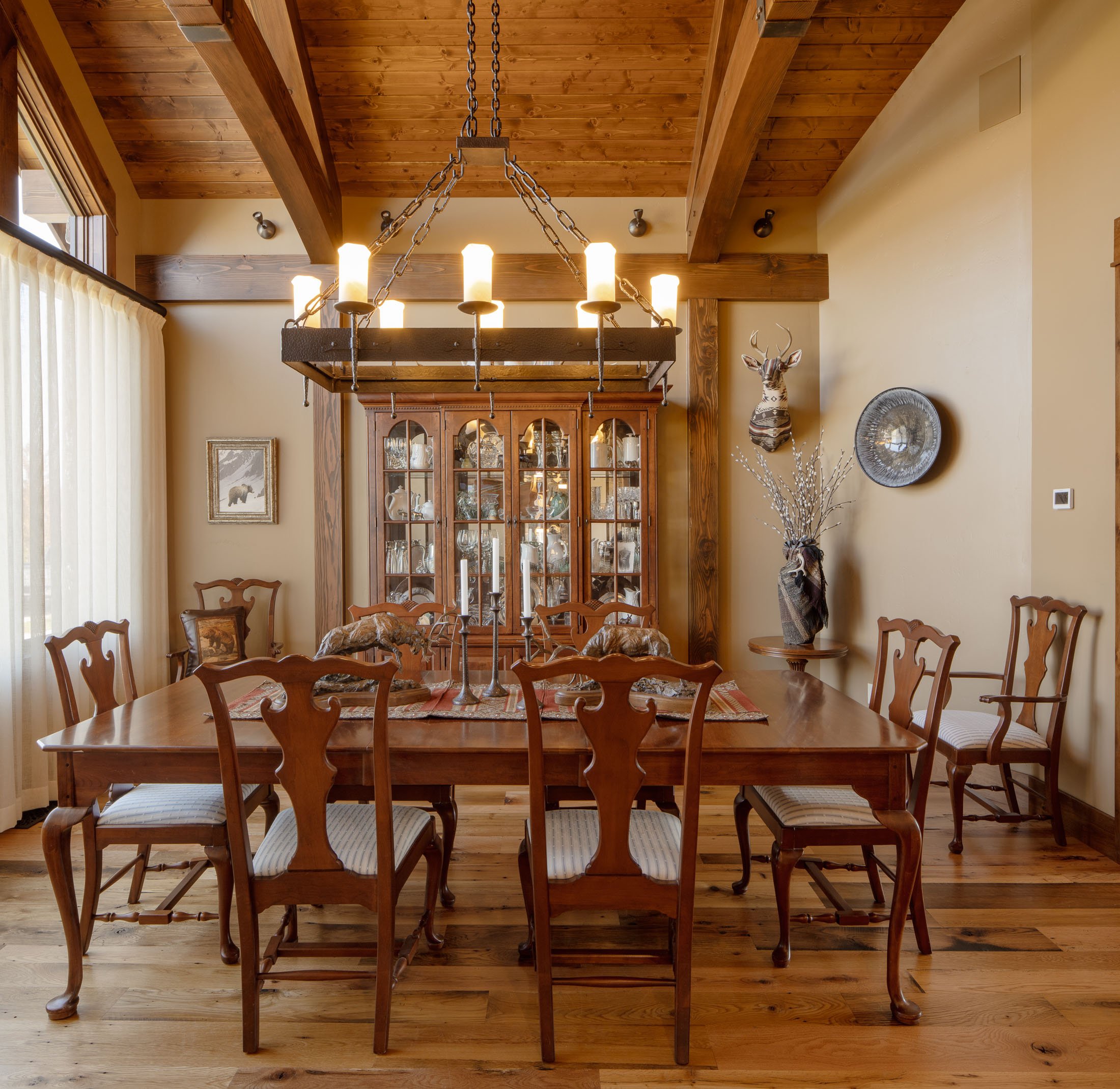 A traditional dining room with a wooden table and eight matching chairs, a large wooden chandelier with candles hanging above, a china cabinet filled with dishes in the background. Custom Home built in Montana by Edgell Building.