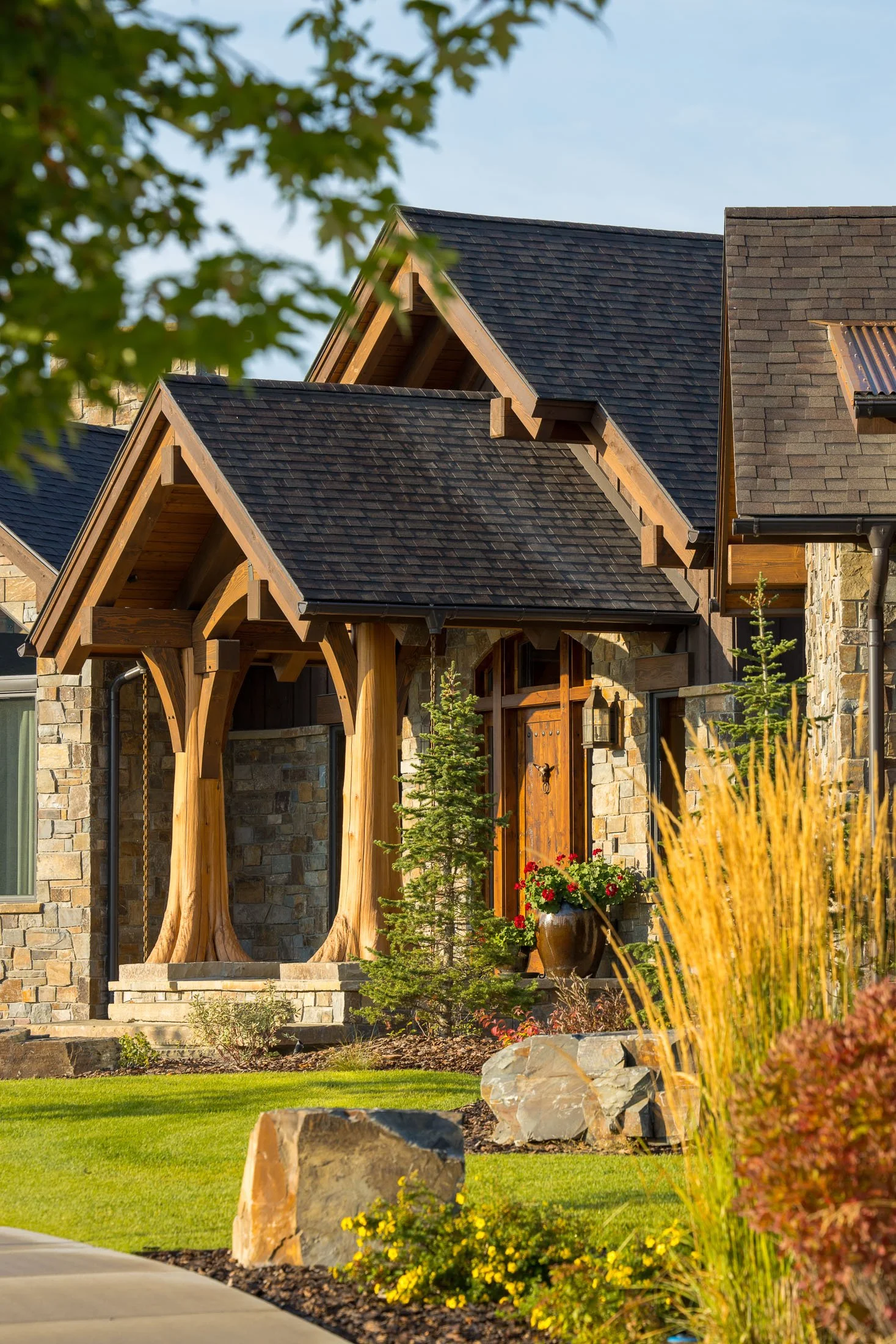 Front view of a house with stone walls, wooden accents, and a variety of plants in the yard, including small trees, yellow and red flowers, and ornamental grasses. Custom Home built in Montana by Edgell Building.