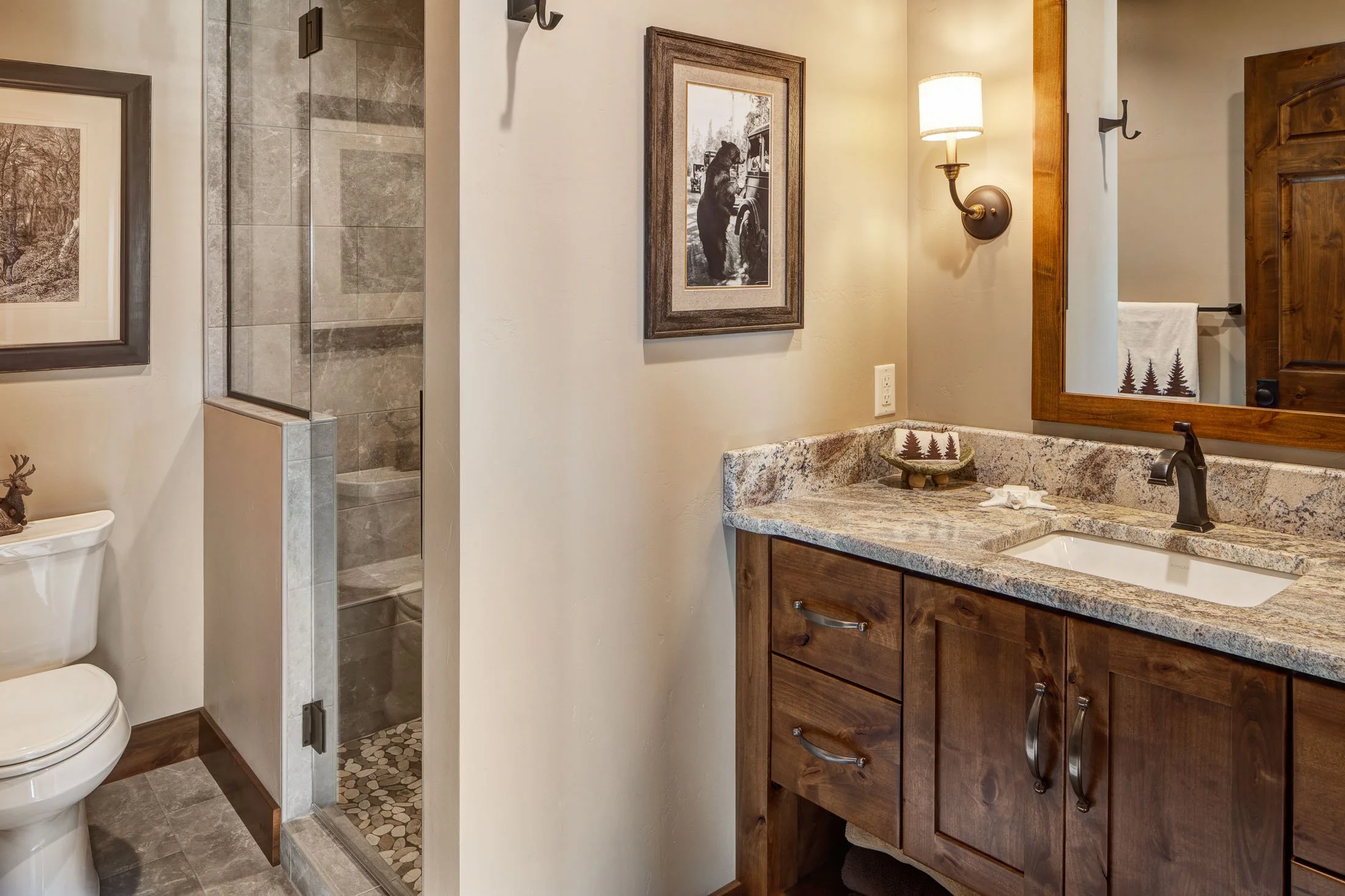 Bathroom with a wooden vanity, granite countertop, mirror, wall sconce, framed photo, and a walk-in shower with tiled walls. Part of a toilet and framed pictures are also visible. Custom Home built in Montana by Edgell Building.