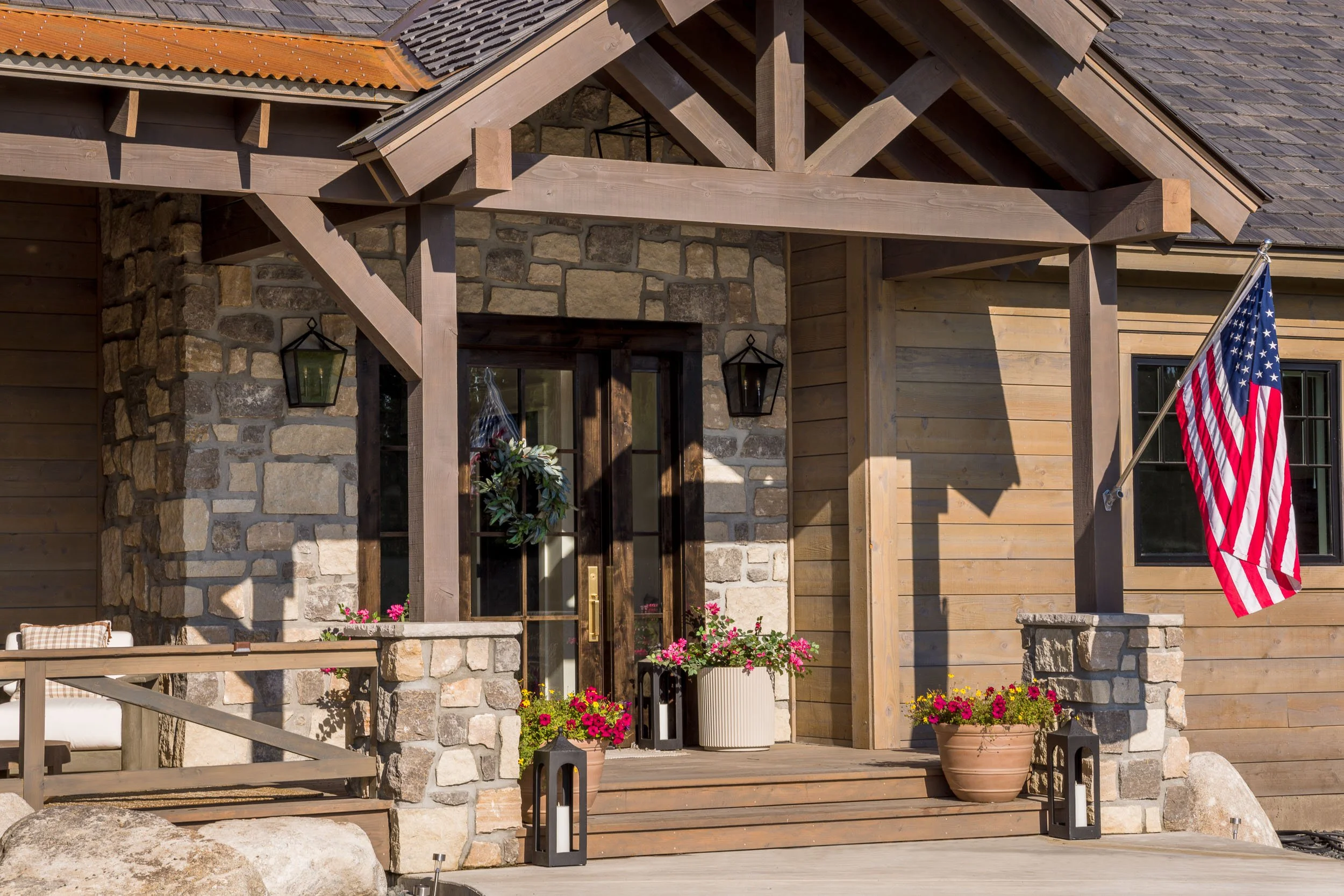 Front porch of a house with stone and wood exterior, decorated with potted flowers and an American flag. Custom Home built in Montana by Edgell Building.