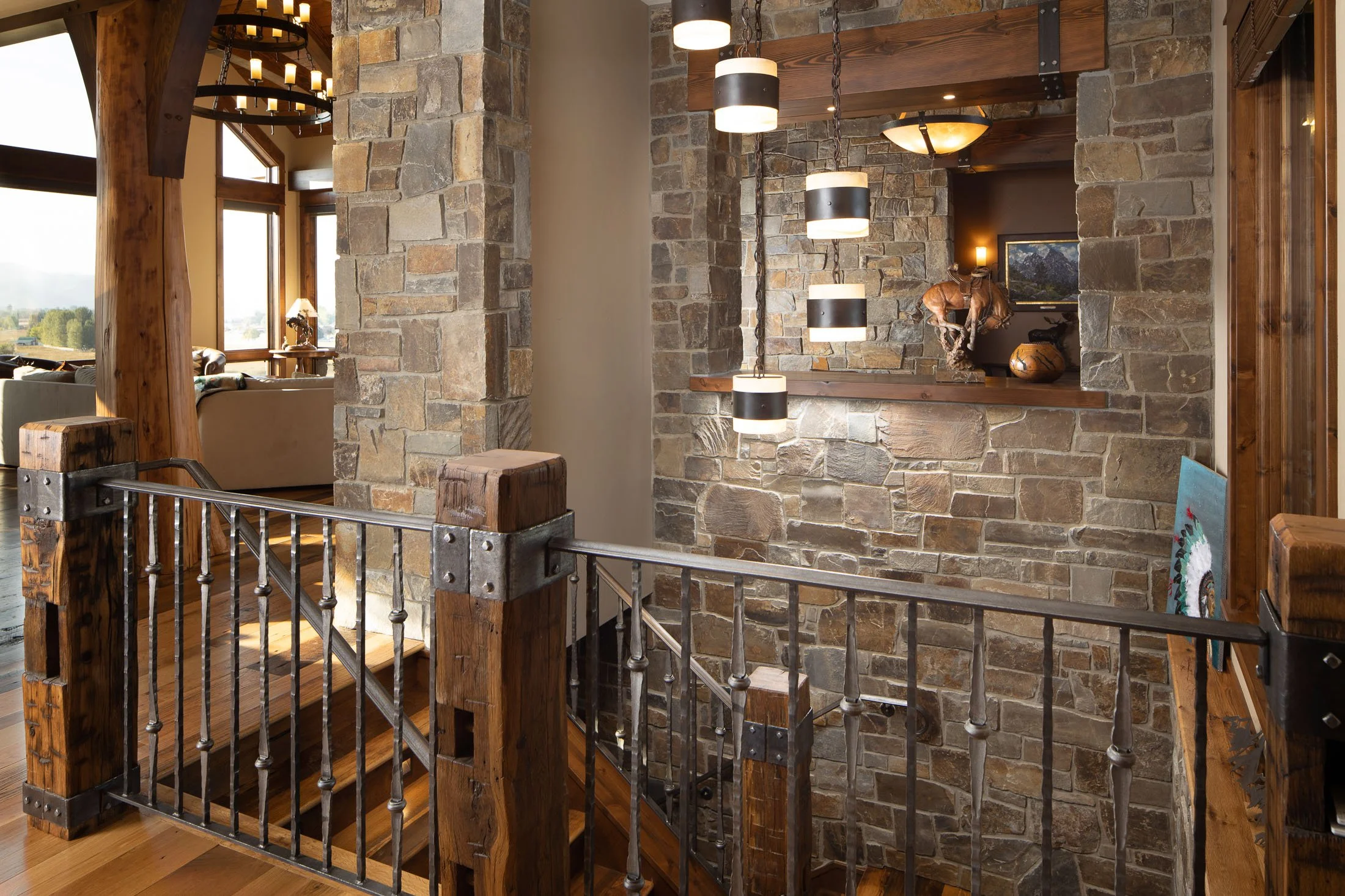 Interior view of a rustic staircase with wooden posts and metal railing, overlooking a living room with large windows and stone walls, decorated with modern and natural elements. Custom Home built in Montana by Edgell Building.