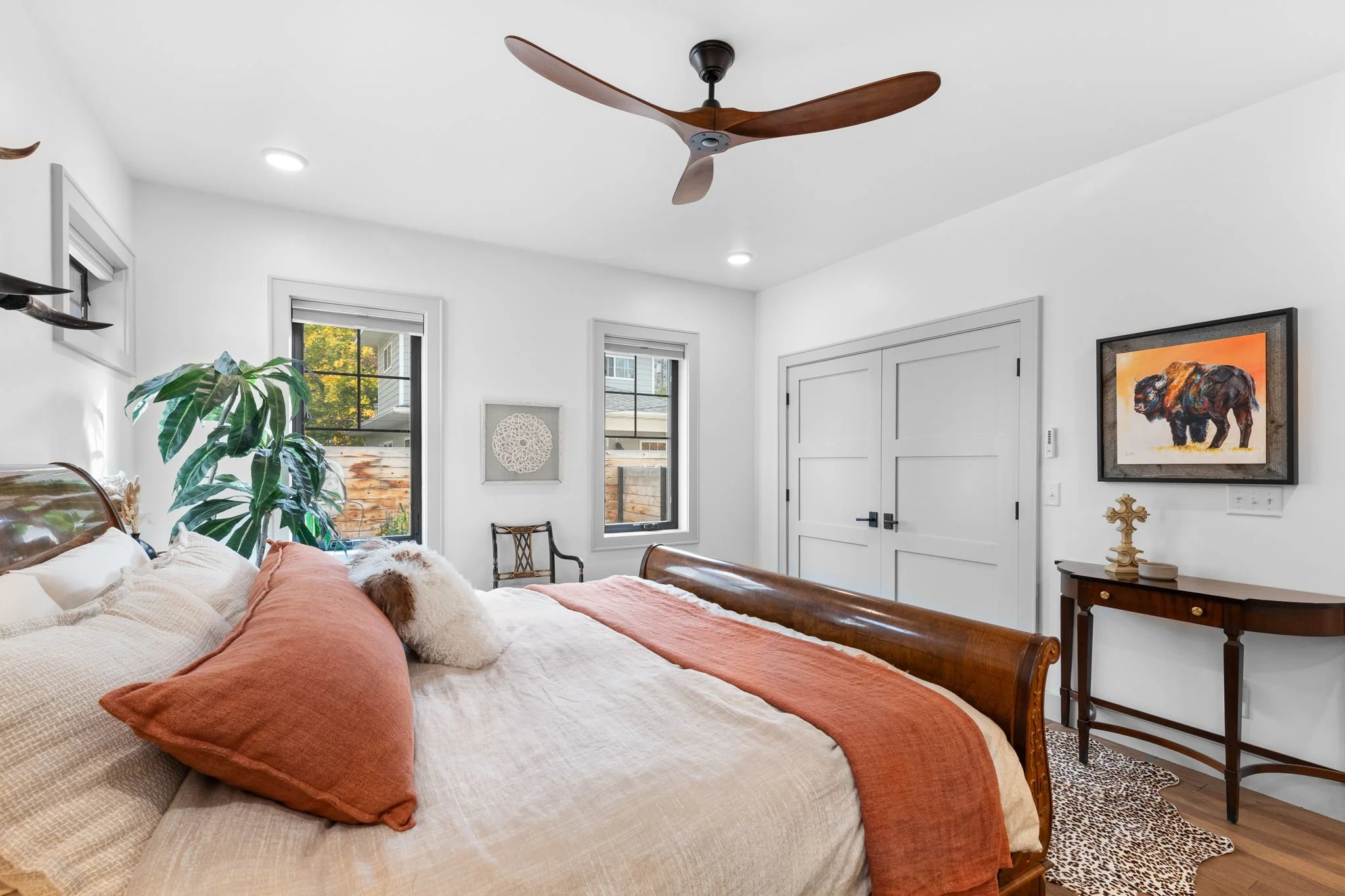 Bedroom with white walls, wooden bed frame, orange and beige bedding, a large plant, two windows, a framed artwork of a bear, and a ceiling fan. Custom Home built in Montana by Edgell Building.