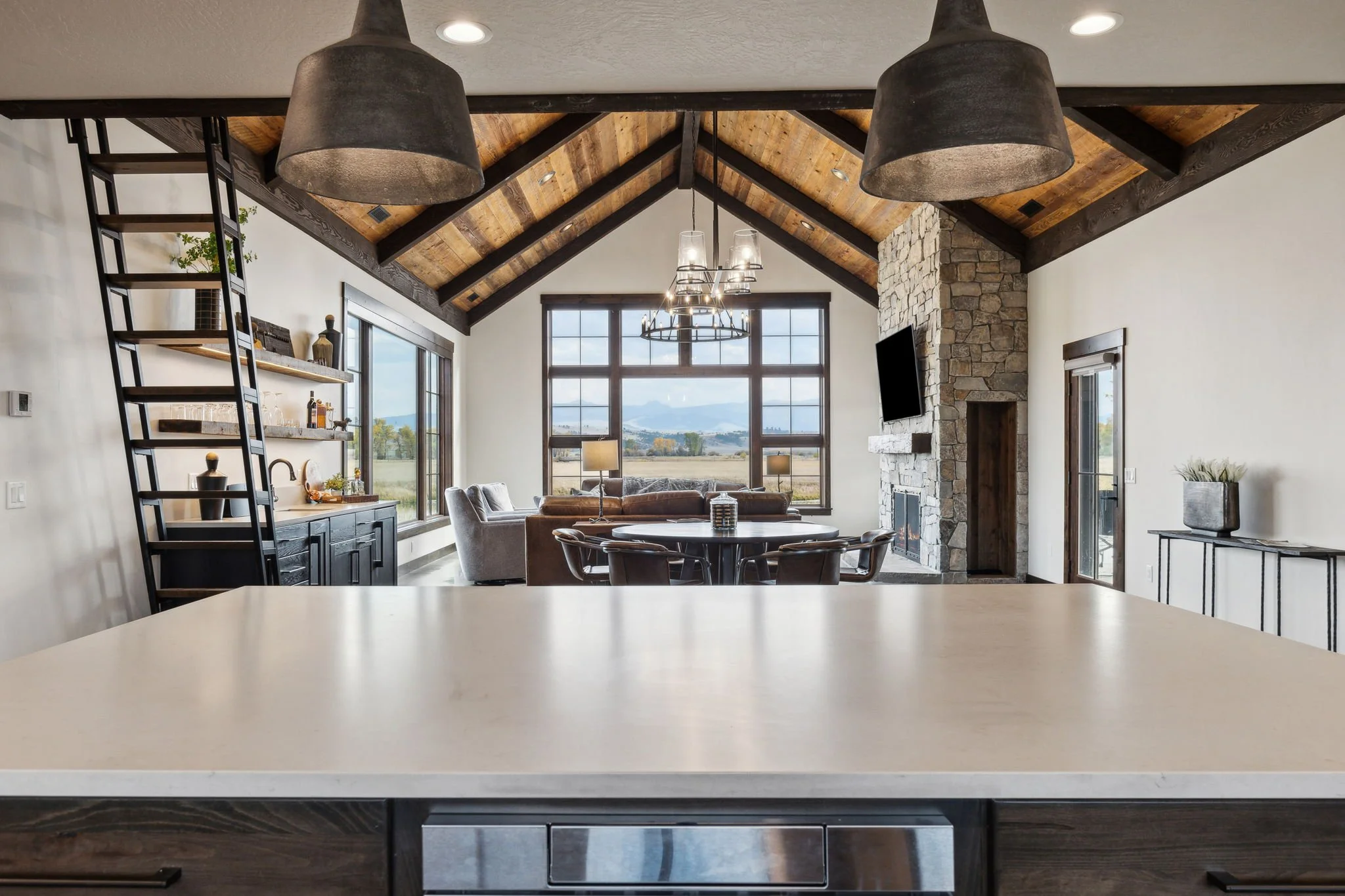 Modern living room with large window, stone fireplace, and vaulted wooden ceiling, viewed from a kitchen island. Custom Home built in Montana by Edgell Building.