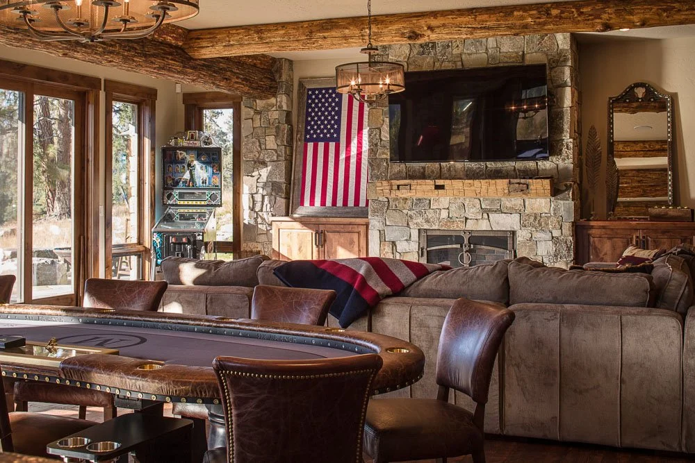 A cozy living room with a stone fireplace, an American flag hanging on the wall, a large TV above the fireplace, a sectional sofa with a striped blanket, a poker table with leather chairs, a slot machine near the corner, large glass doors leading out