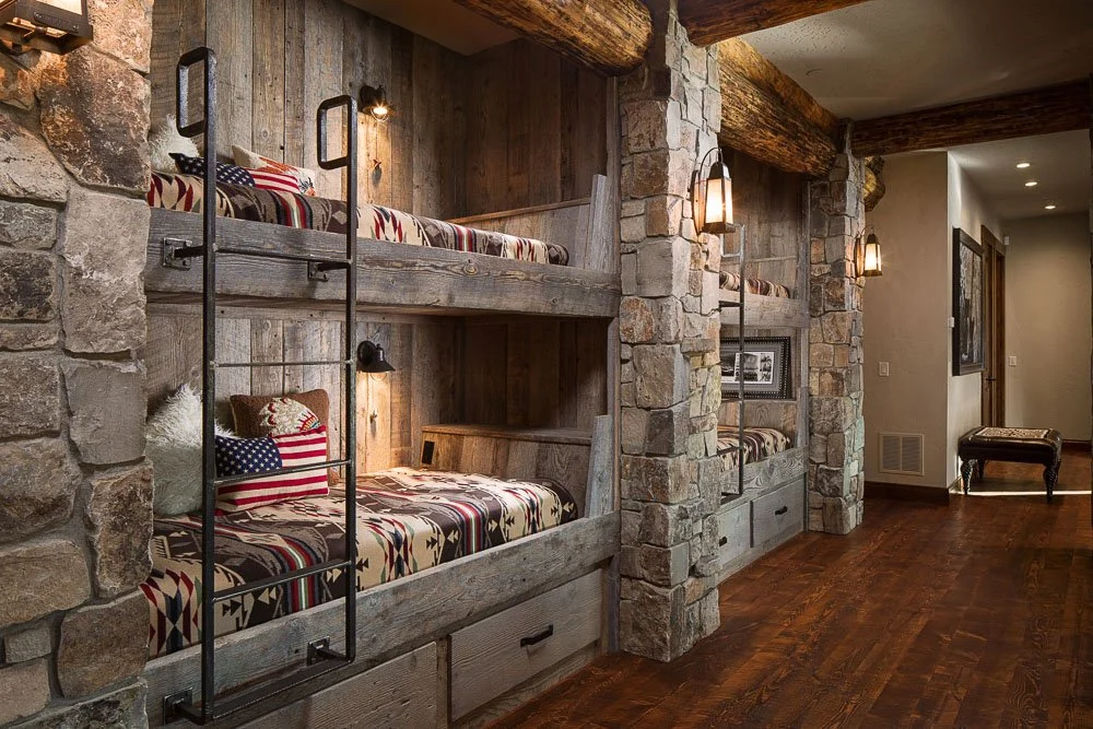 Interior of a rustic cabin room with bunk beds built into stone and wood walls, decorated with American flag-themed bedding and pillows, warm lighting fixtures, and wooden beams. Custom Home built in Montana by Edgell Building.