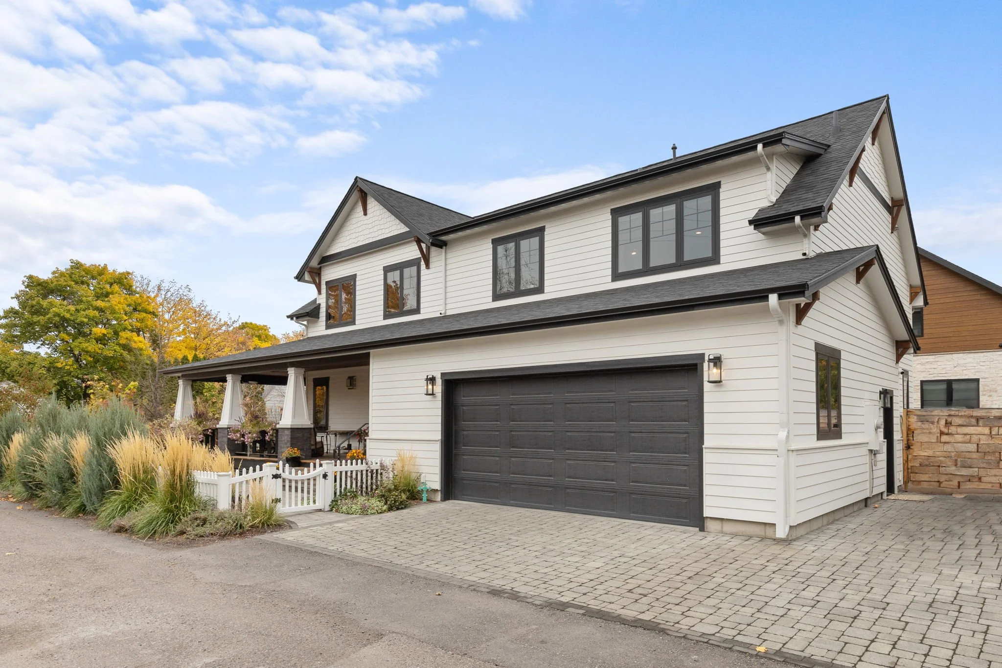 Front view of a modern two-story white house with black garage door and trim, black window frames, and a small porch with white railings, surrounded by landscaping and trees with fall foliage. Custom Home built in Montana by Edgell Building.