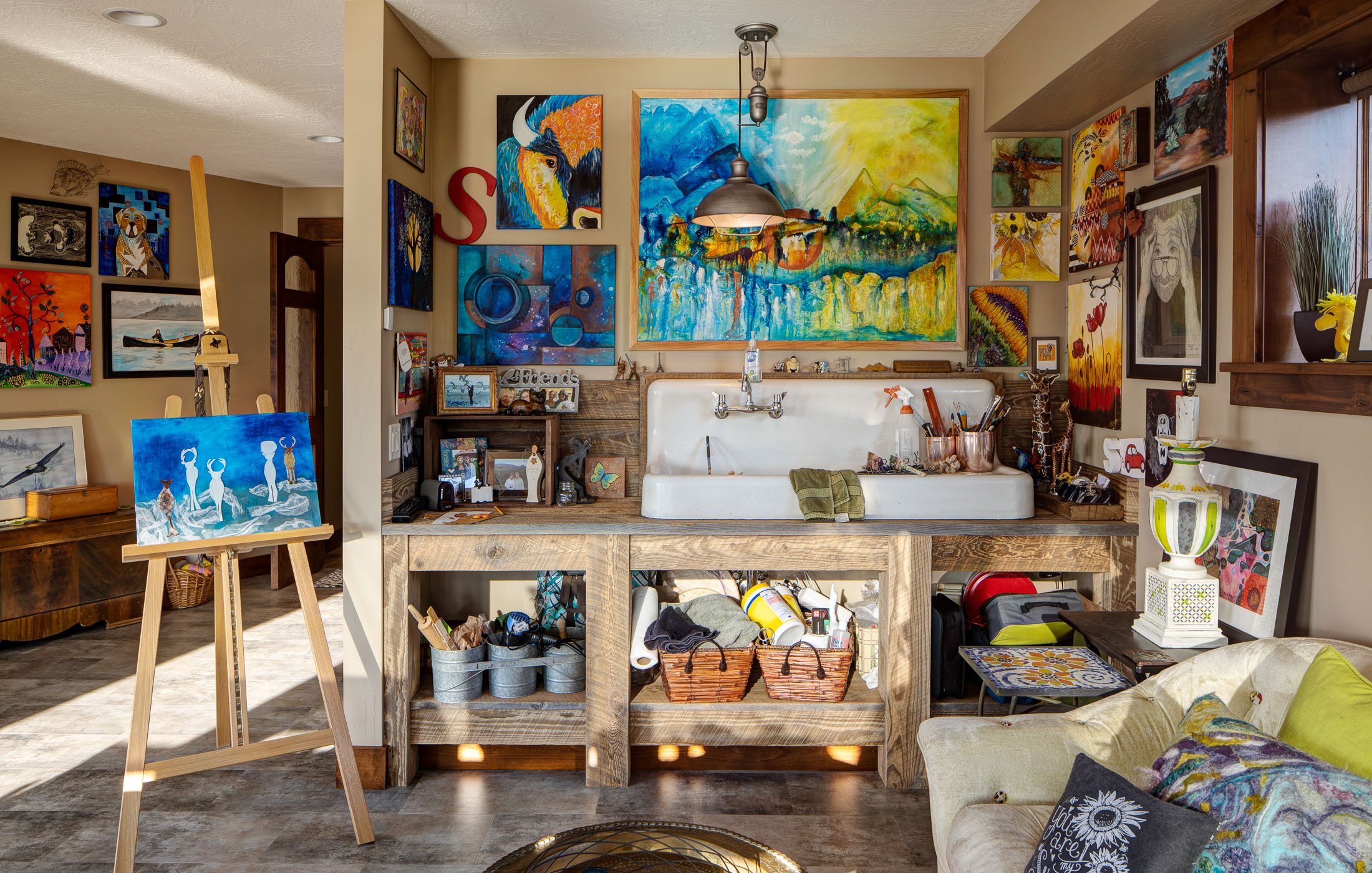 Interior of an art studio with colorful paintings on walls, a large sink with art supplies, an easel with a painting, and a couch with pillows. Custom Home built in Montana by Edgell Building.