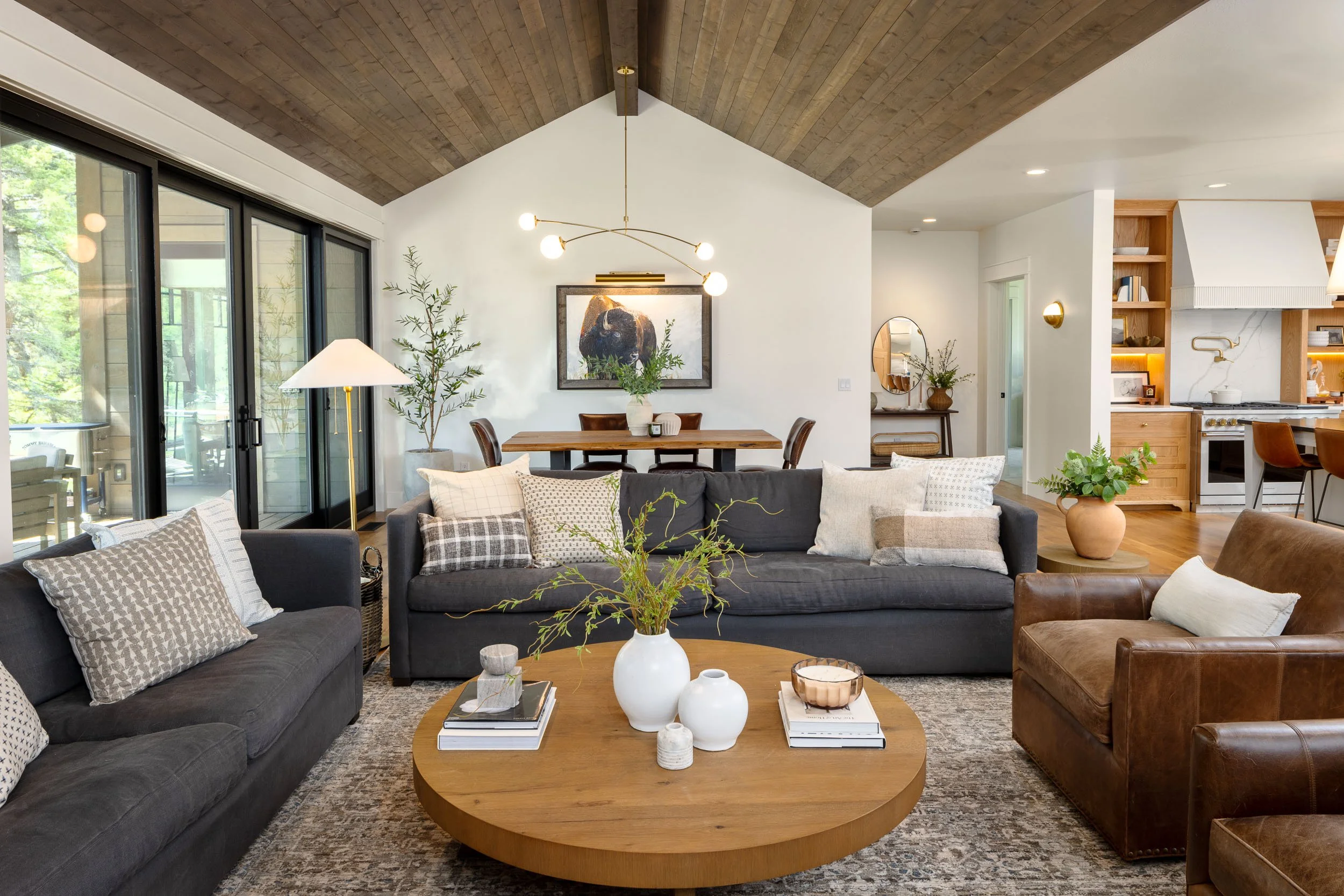 Living room with large gray sofa, brown leather armchair, round wooden coffee table with vases, and decorative pillows, with large glass sliding doors leading to a patio, and a wooden dining table and chairs in the background, under a wood-paneled ce