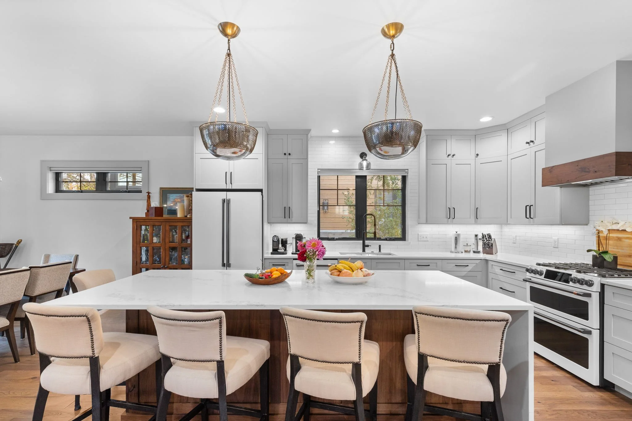 Modern kitchen with white cabinets, a large island with beige chairs, and two pendant lights hanging from the ceiling. Custom Home built in Montana by Edgell Building.