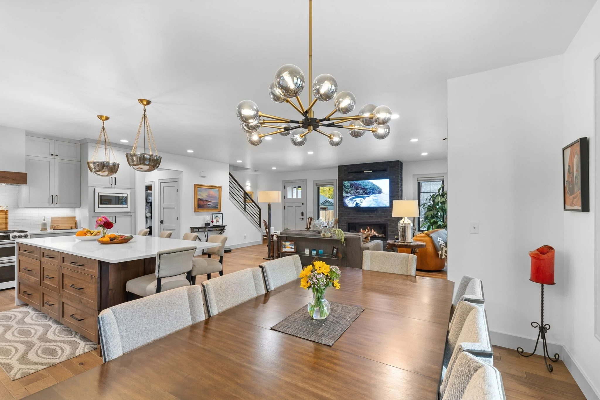 Open concept living and dining room with a wooden dining table, a modern chandelier, a kitchen island, and a cozy fireplace with a television above. Custom Home built in Montana by Edgell Building.