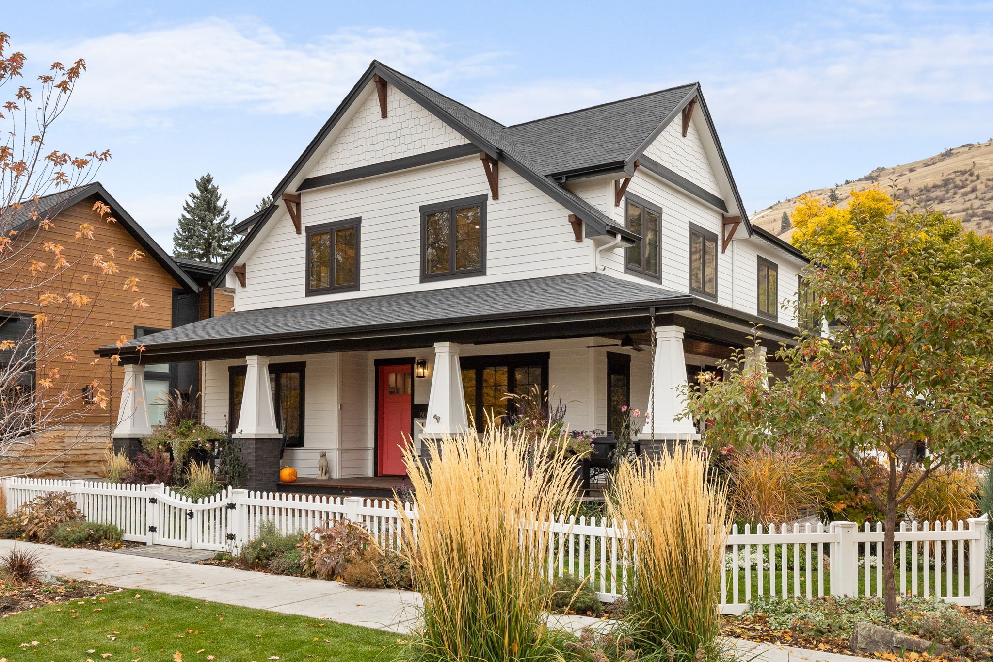 A modern two-story house with white siding, black trim, and a red front door, surrounded by a white picket fence, autumn trees, and landscaped front yard. Custom Home built in Montana by Edgell Building.