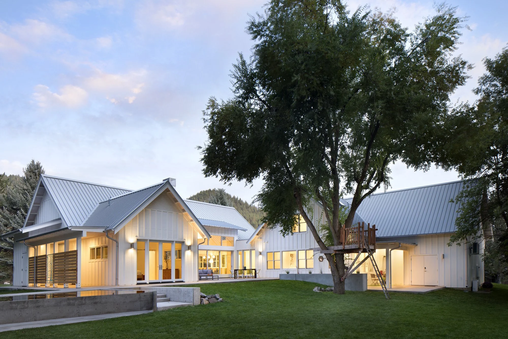 Modern white house with large windows and a metal roof, lit from inside, with a large tree in the front yard and a mountain in the background during dusk. Custom Home built in Montana by Edgell Building.
