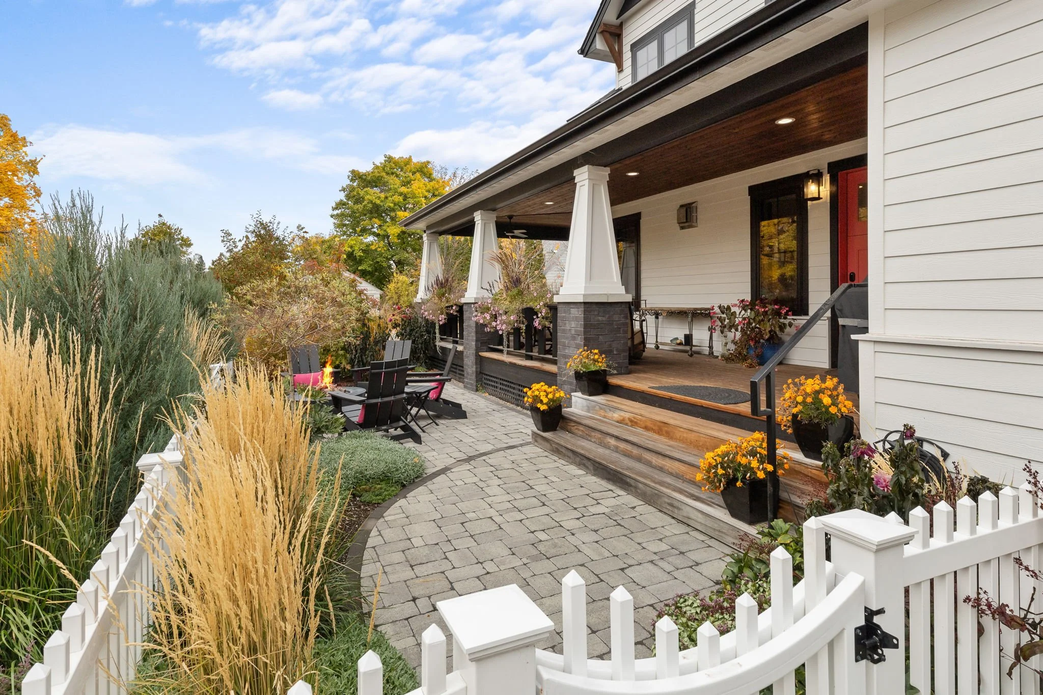 A cozy backyard patio with a stone pathway, a fire pit, black Adirondack chairs with pink cushions, colorful potted flowers, and a porch with seating, surrounded by greenery and trees. Custom Home built in Montana by Edgell Building.