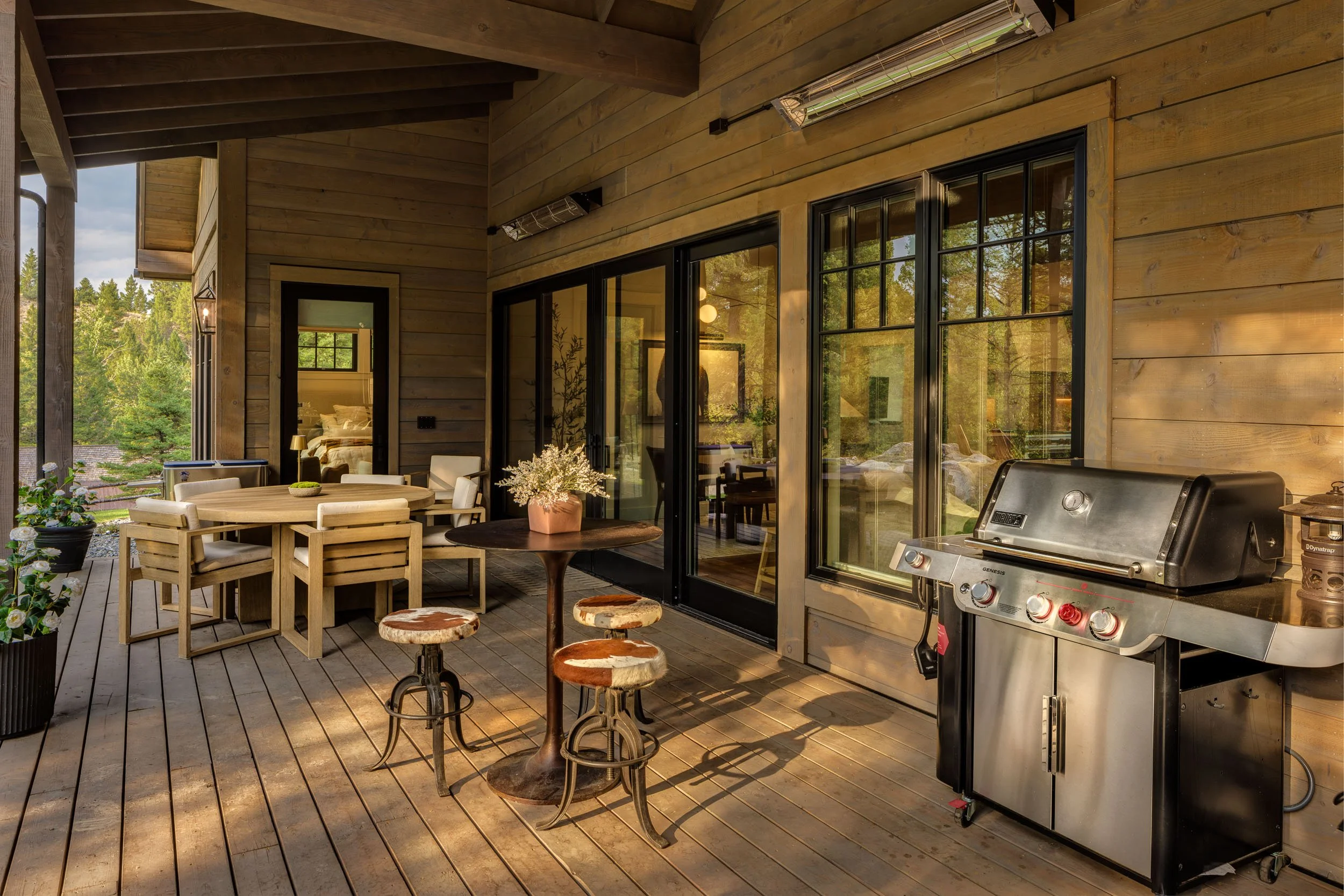 Wooden deck with patio furniture, potted plants, a gas grill, and sliding glass doors leading into a house, overlooking greenery. Home Designed and Built By Edgell Building in Western Montana.