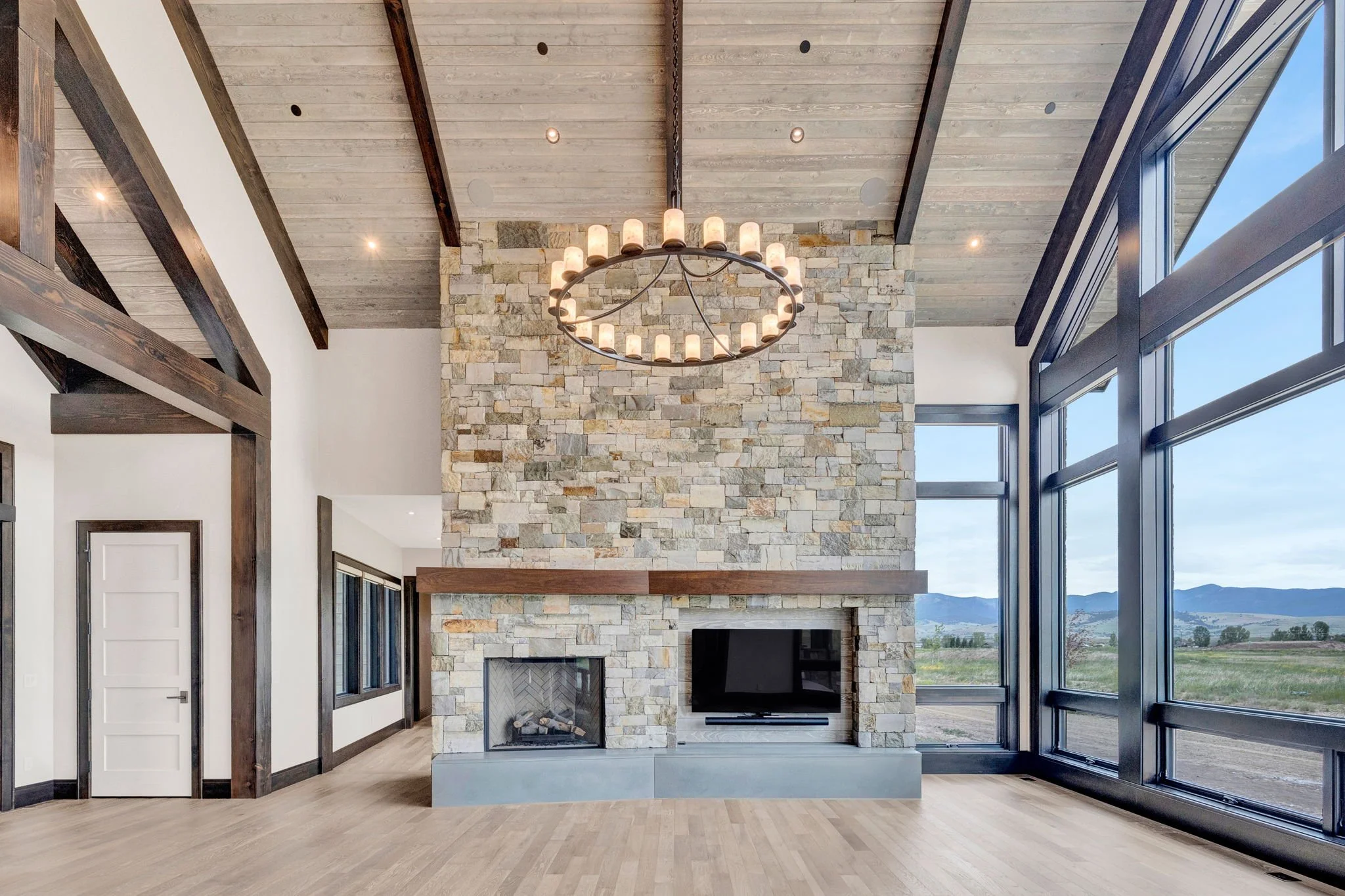 Interior of a modern living room with a large stone fireplace, a mounted TV, wooden ceiling beams, a chandelier, and large windows showing a scenic mountain view. Custom Home built in Montana by Edgell Building.