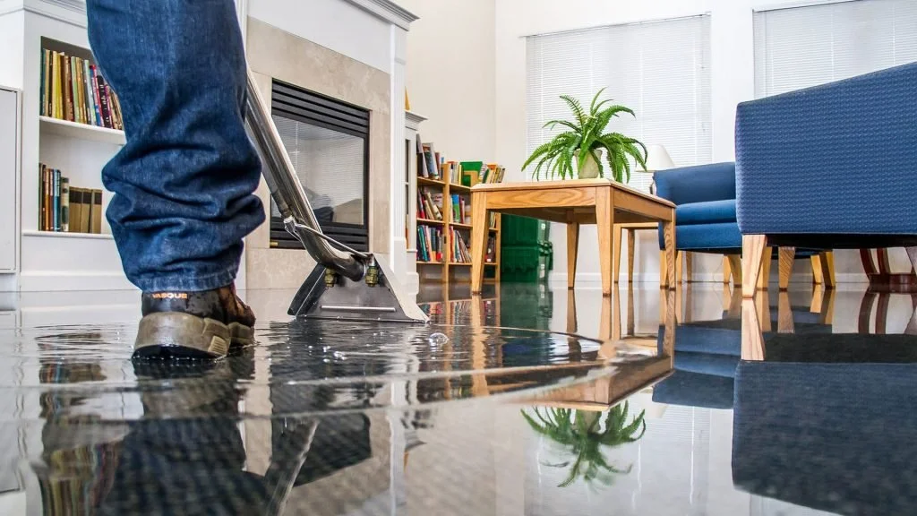 Person using a squeegee to clean up a large water spill on the floor in a living room with bookshelves, a fireplace, a wooden coffee table, and green plants.
