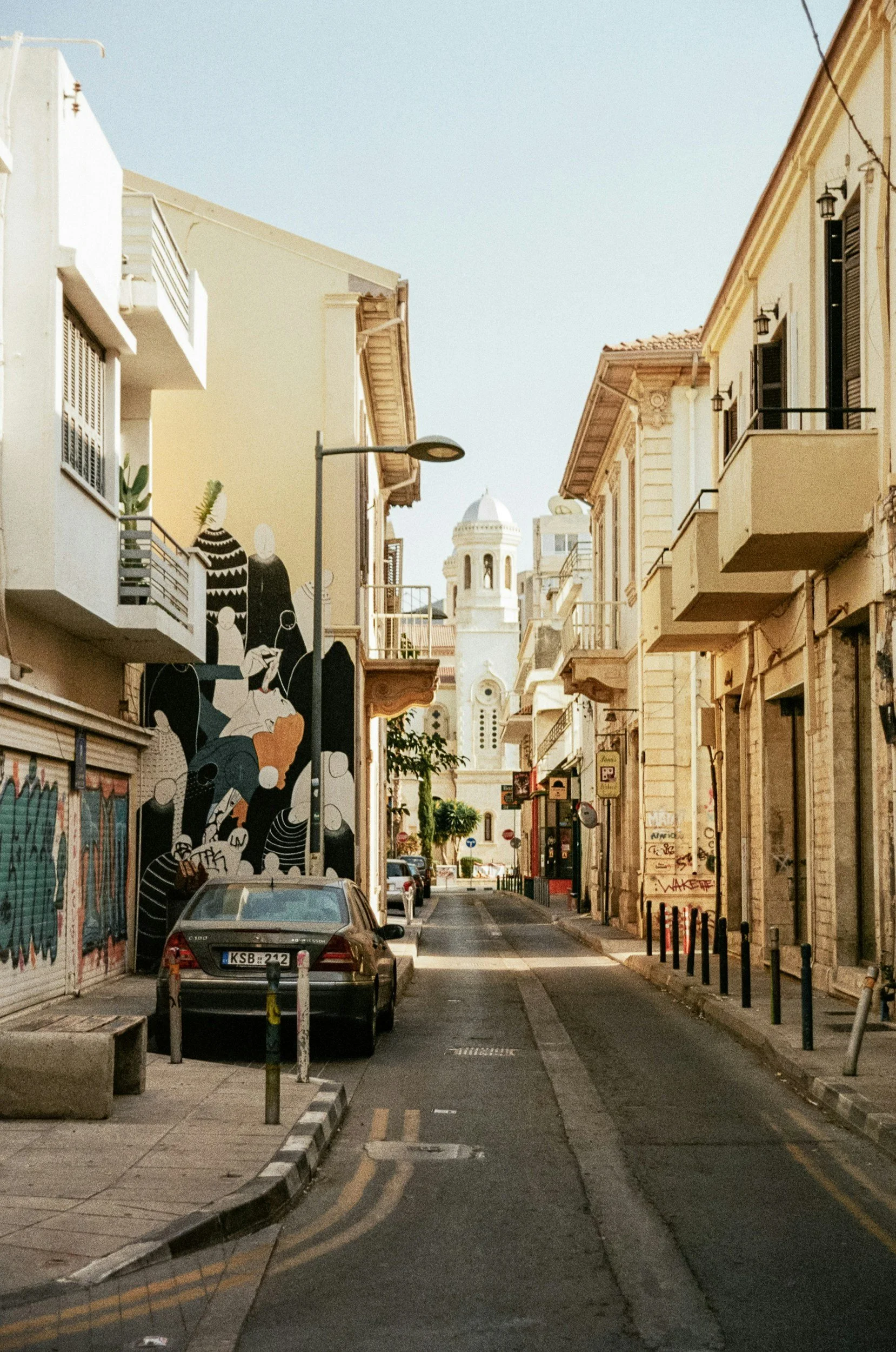 A narrow city street with parked cars, buildings with balconies, and a church tower in the background.
