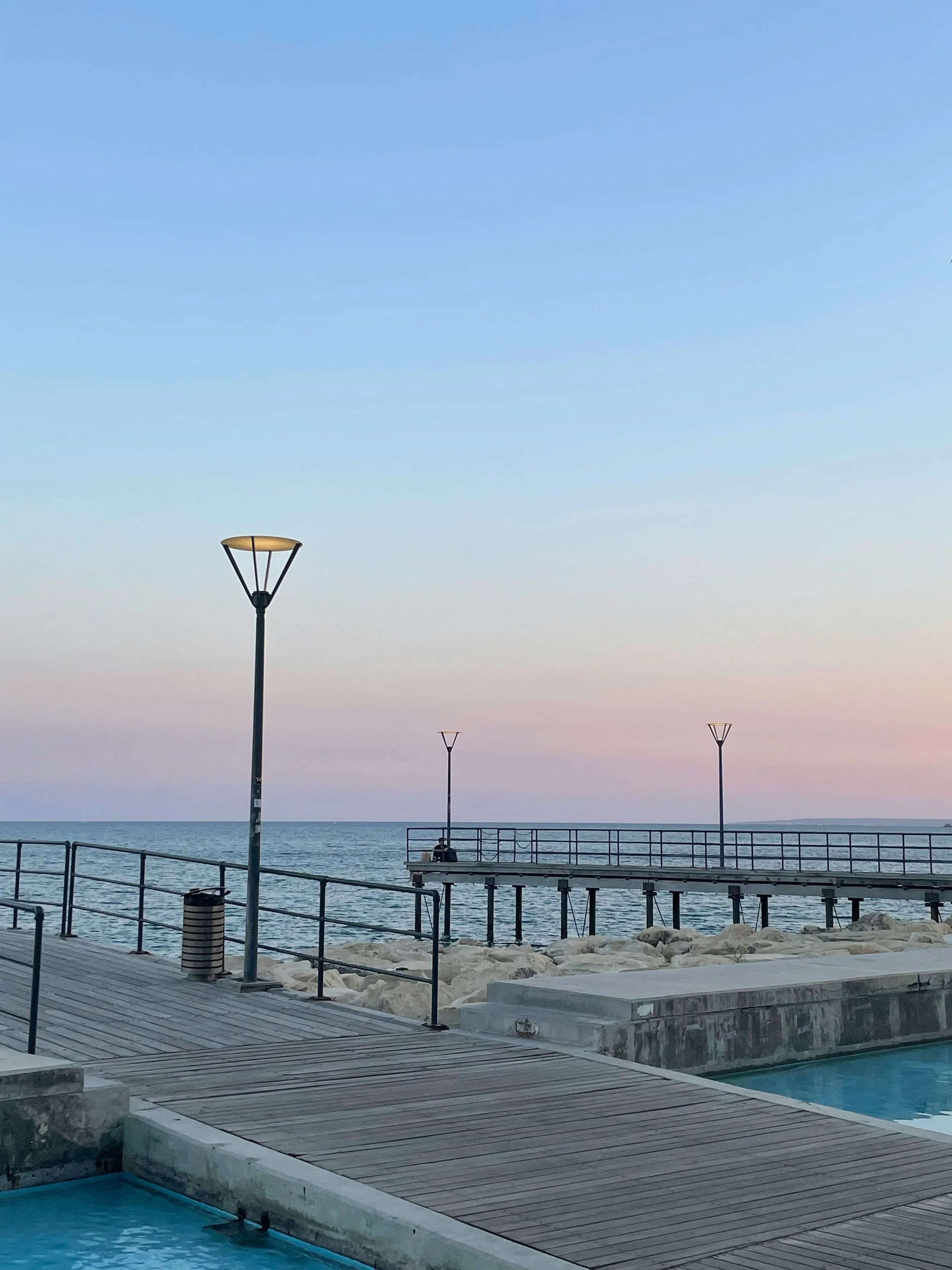 A seaside scene at dusk or dawn, featuring a wooden dock with metal railings extending over rocks into the water, three lamp posts, and a clear sky with a gentle gradient of pastel colors.