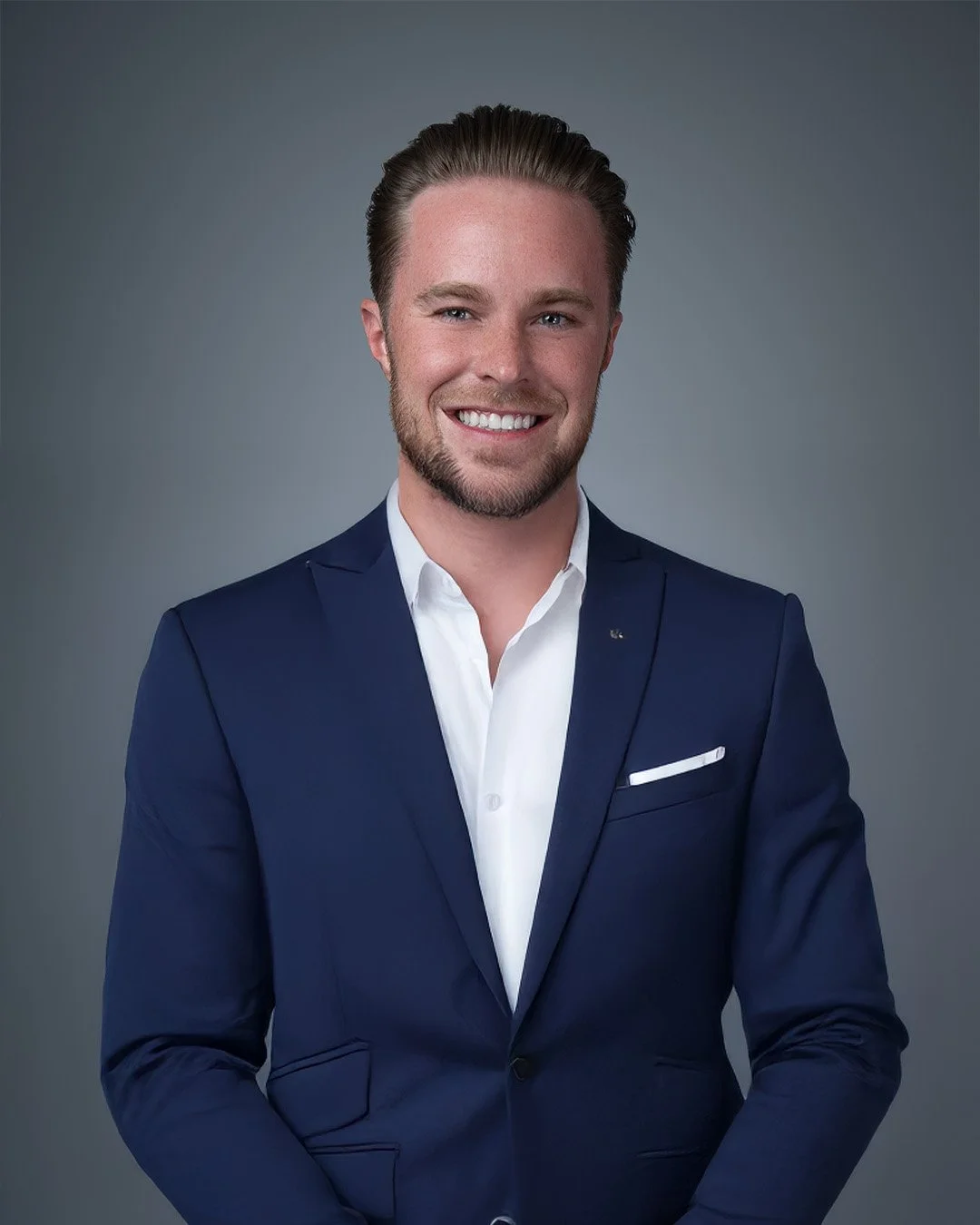 Sam Northern Nevada Realestate Professinoal A professional man with short brown hair, a beard, and blue eyes, wearing a navy blue suit jacket, a white dress shirt, and a white pocket square, smiling against a gray background.