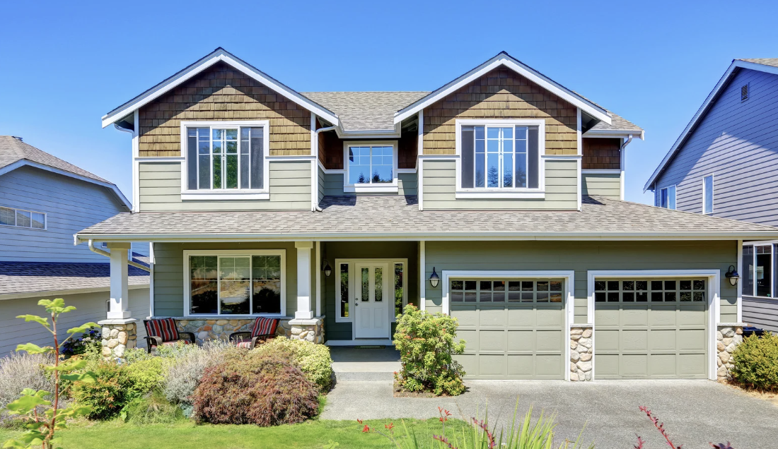 Front view of a two-story suburban house with green siding and stone accents, featuring a large driveway, landscaped yard, and two-car garage.
