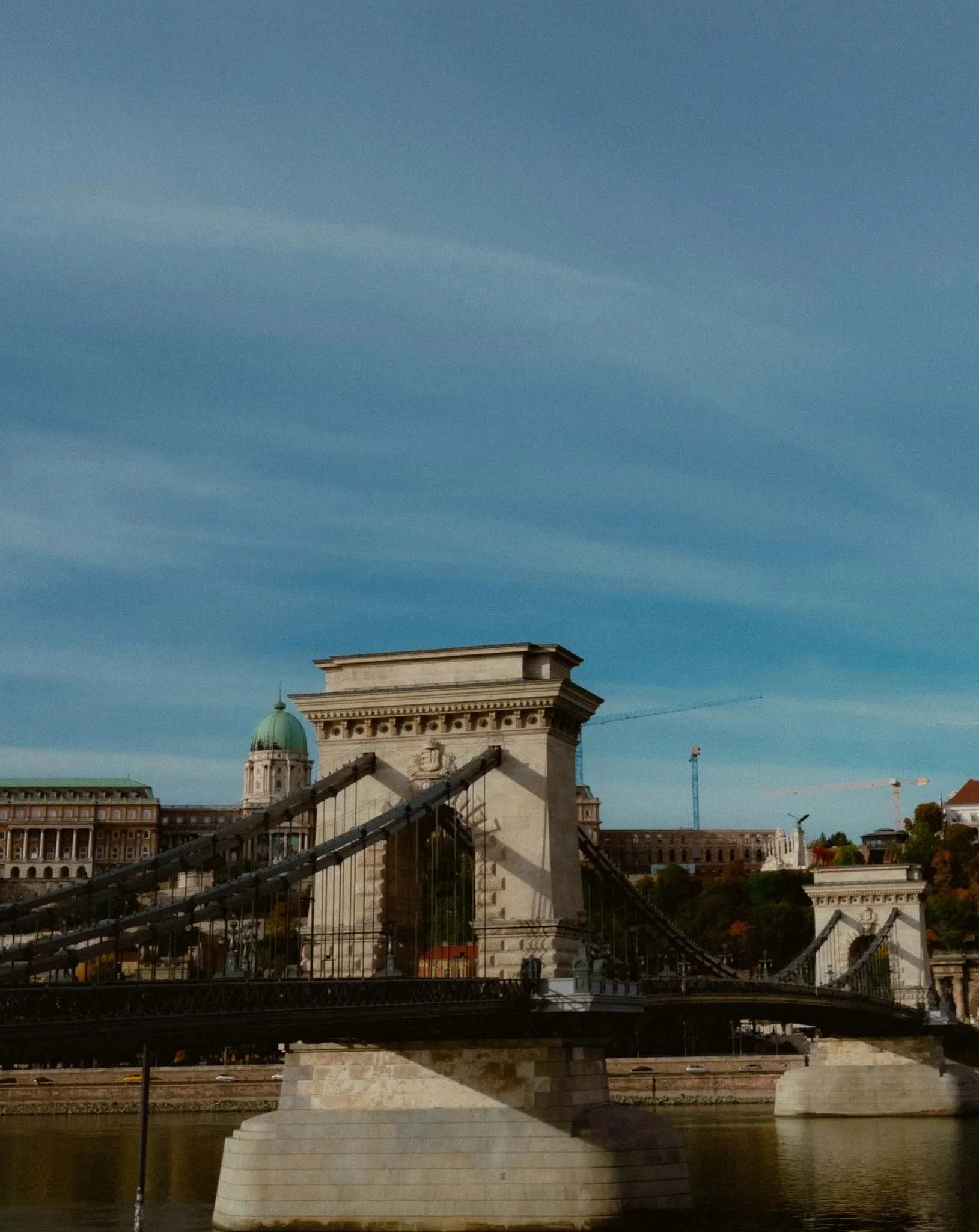 View of the Chain Bridge over the Danube River in Budapest with historic buildings and construction cranes in the background under a clear blue sky.