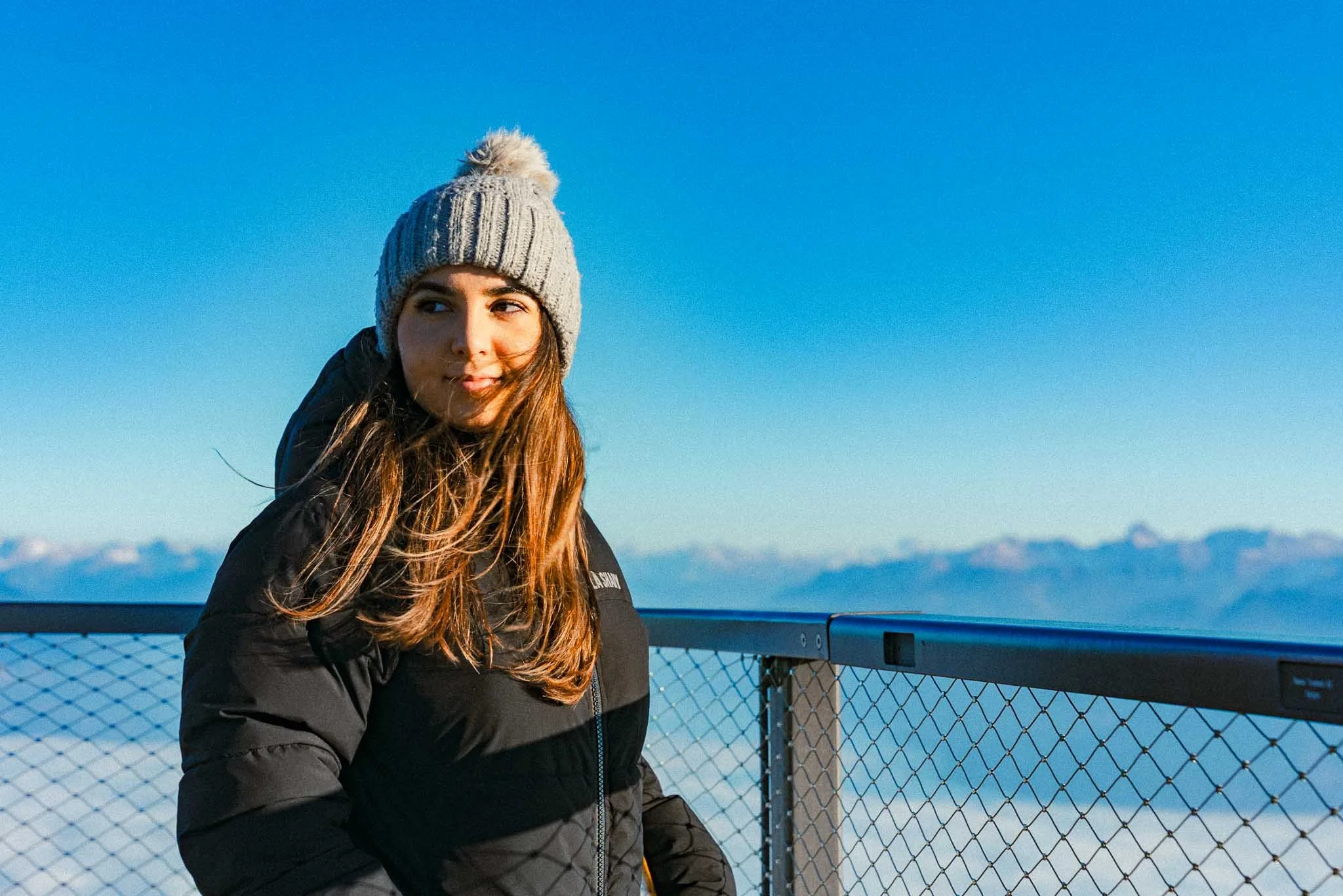 A young woman with long brown hair wearing a gray knit beanie with a pom-pom and a black winter jacket standing on a lookout point with a metal railing, overlooking snow-capped mountains under a clear blue sky.