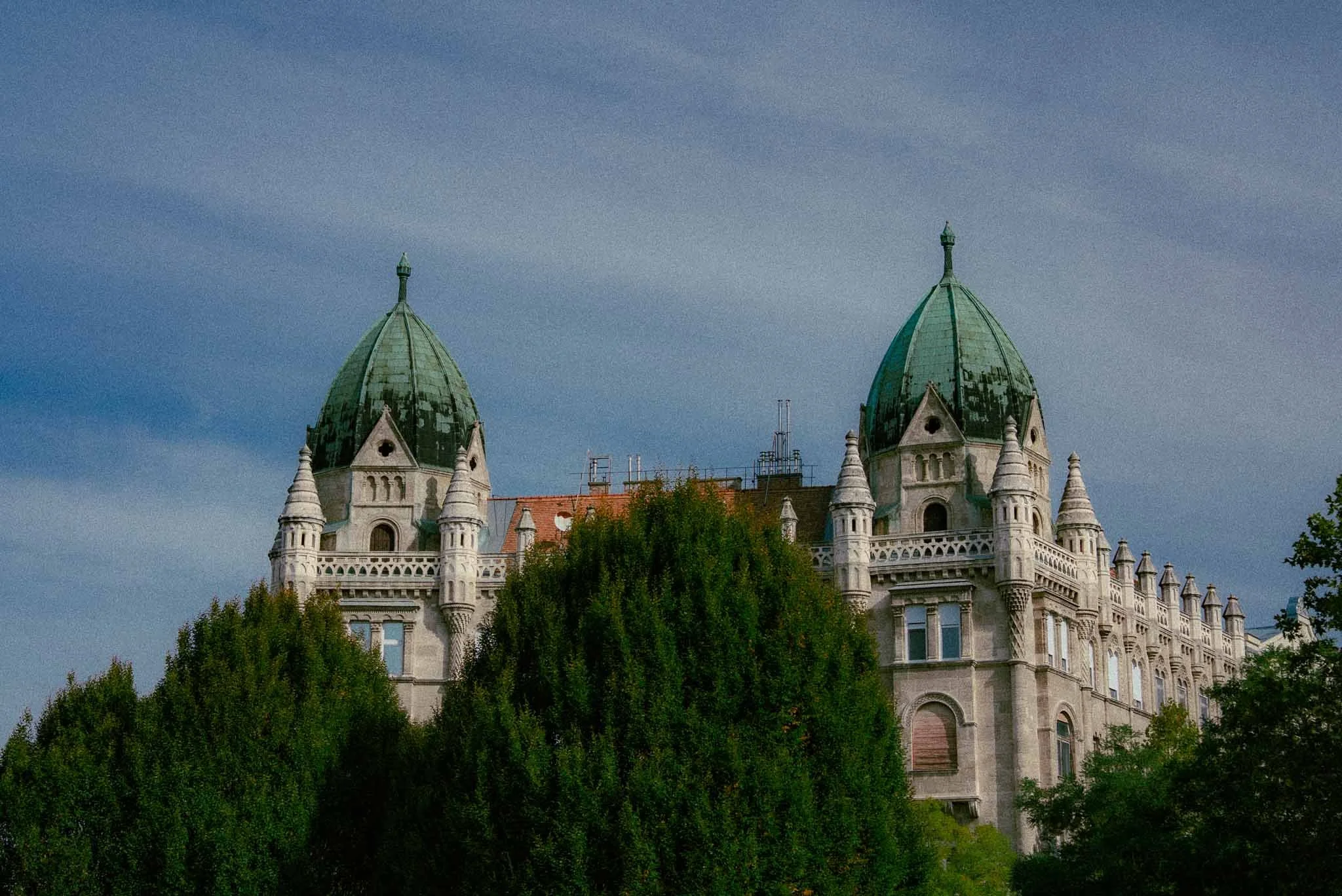A castle with two green domed towers, ornate architecture, surrounded by green trees under a cloudy sky.