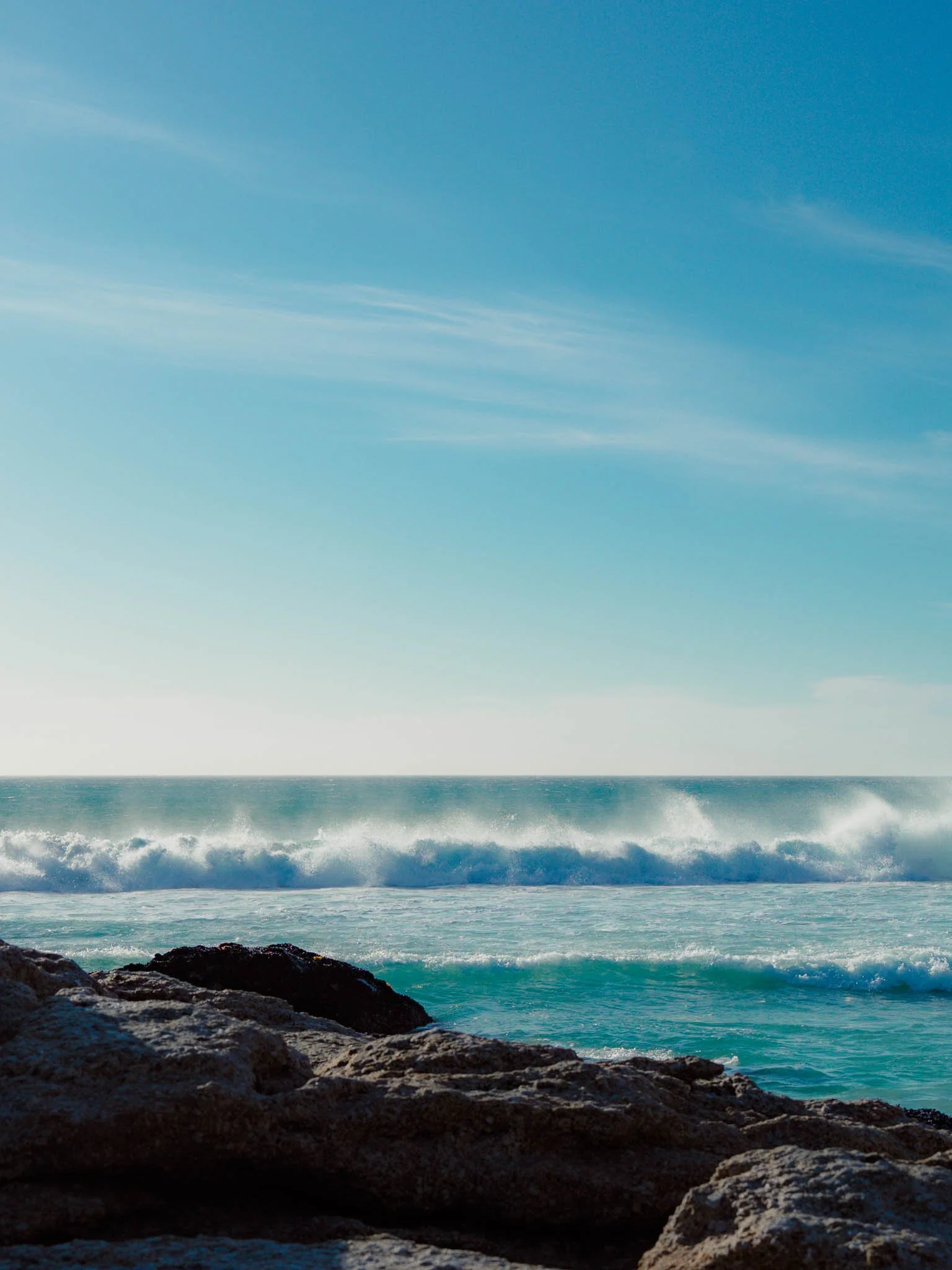 A view of the ocean with waves crashing against rocks in the foreground, blue sky with wispy clouds above.