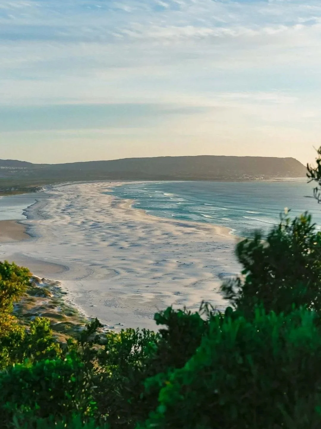 A coastal view of a beach with waves, sandy shore, and green foliage in the foreground, with a distant landmass and cloudy sky in the background.
