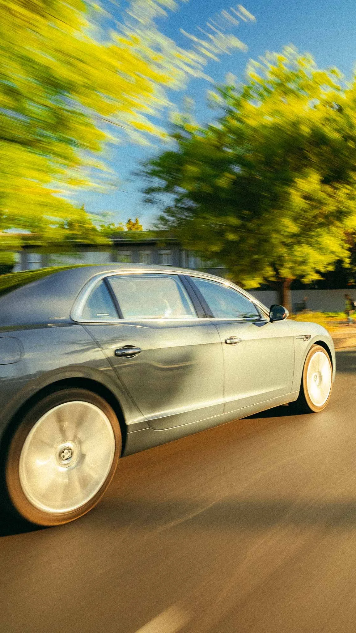 A silver luxury sedan driving on a tree-lined street with green foliage and houses in the background, captured in motion blur.