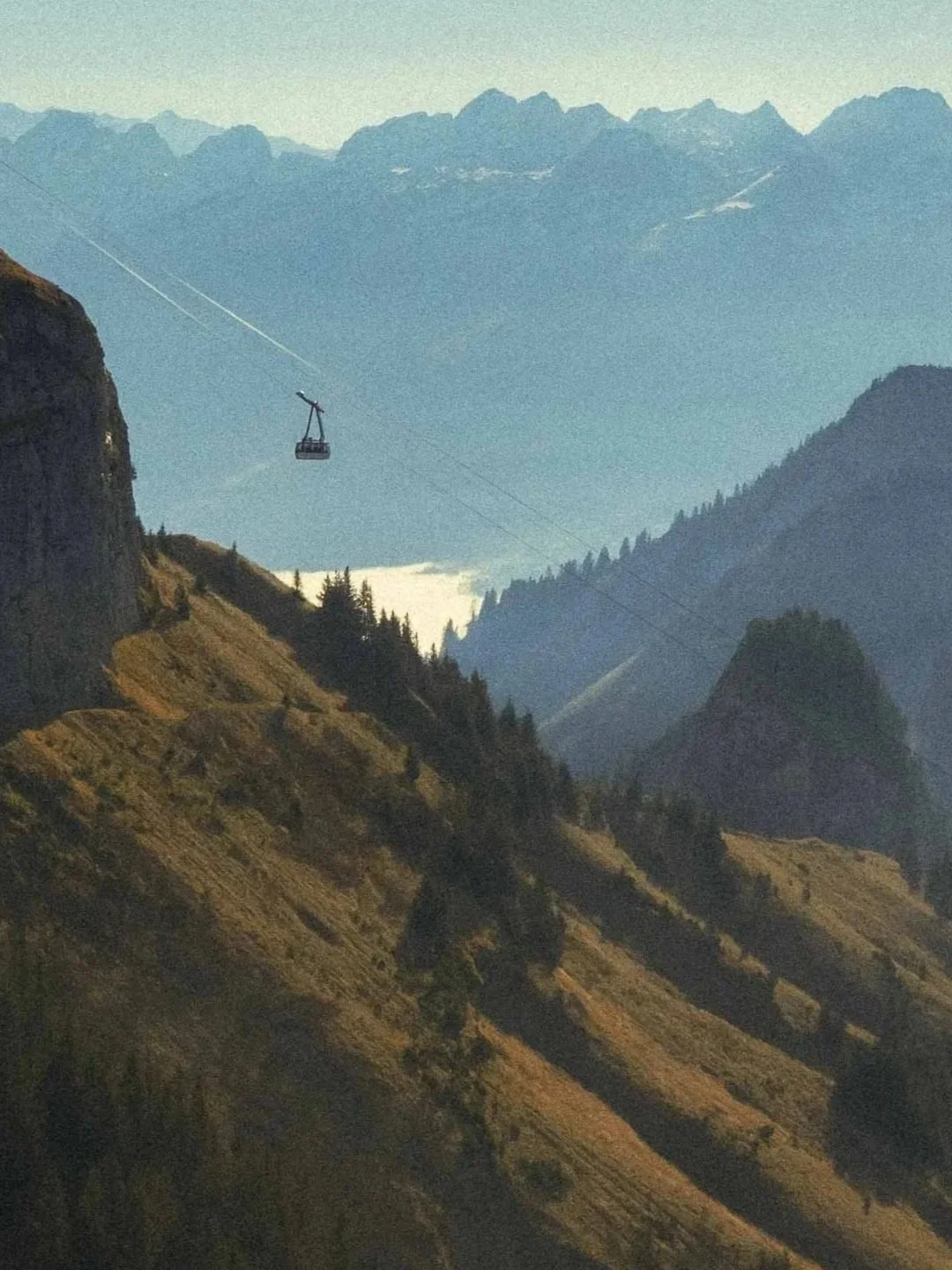 A mountain landscape with rugged terrain and pine trees, with a ski lift cable running across the scene and a single ski lift chair hanging in the air, against a backdrop of towering snow-capped peaks.