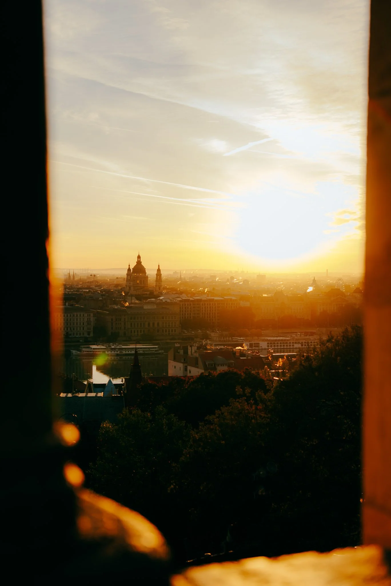 Sunrise over a city skyline viewed through a window, with buildings and a river visible. The image has warm golden hues.