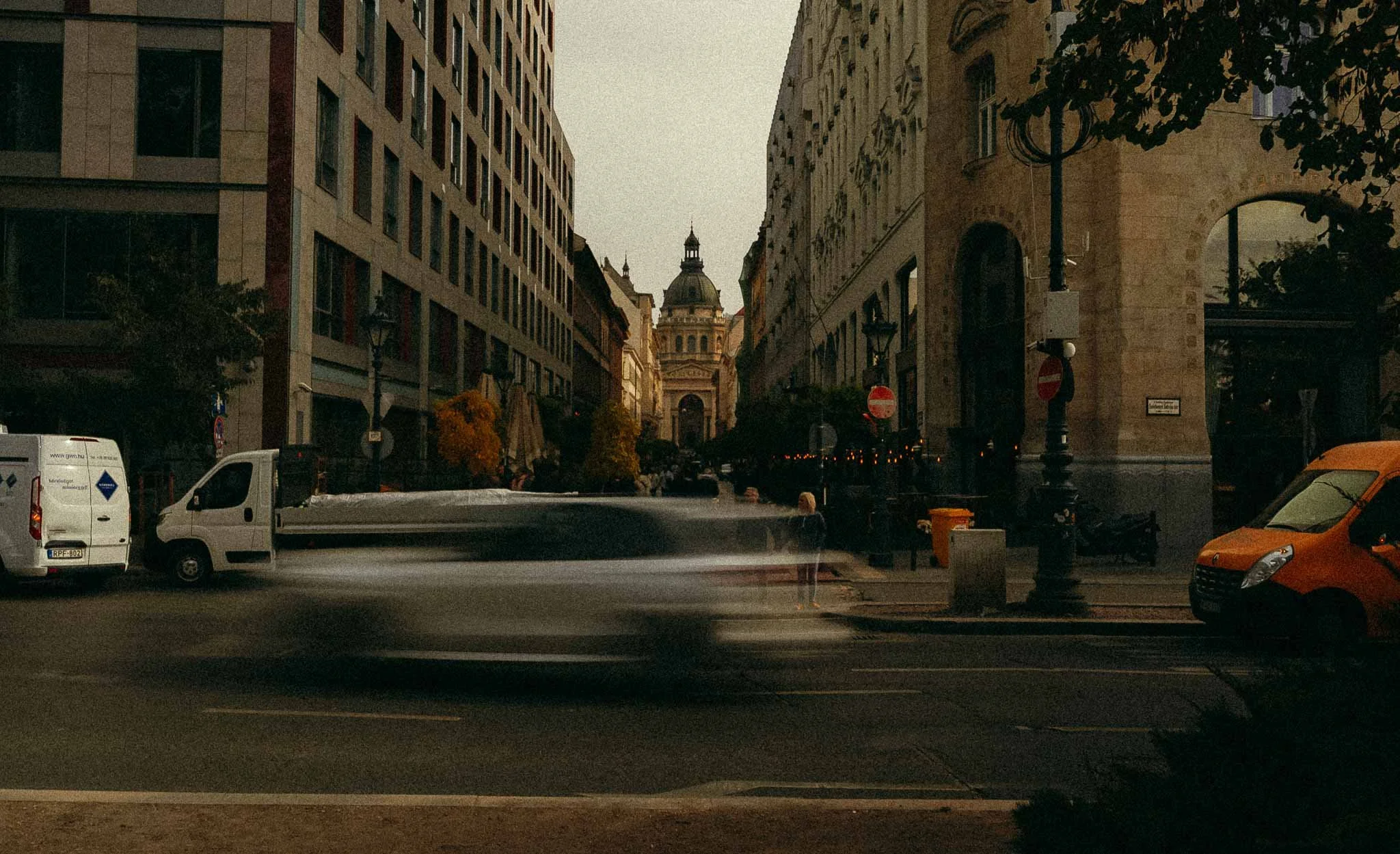 A street view with cars passing by, buildings on both sides, and a domed historical building in the background.