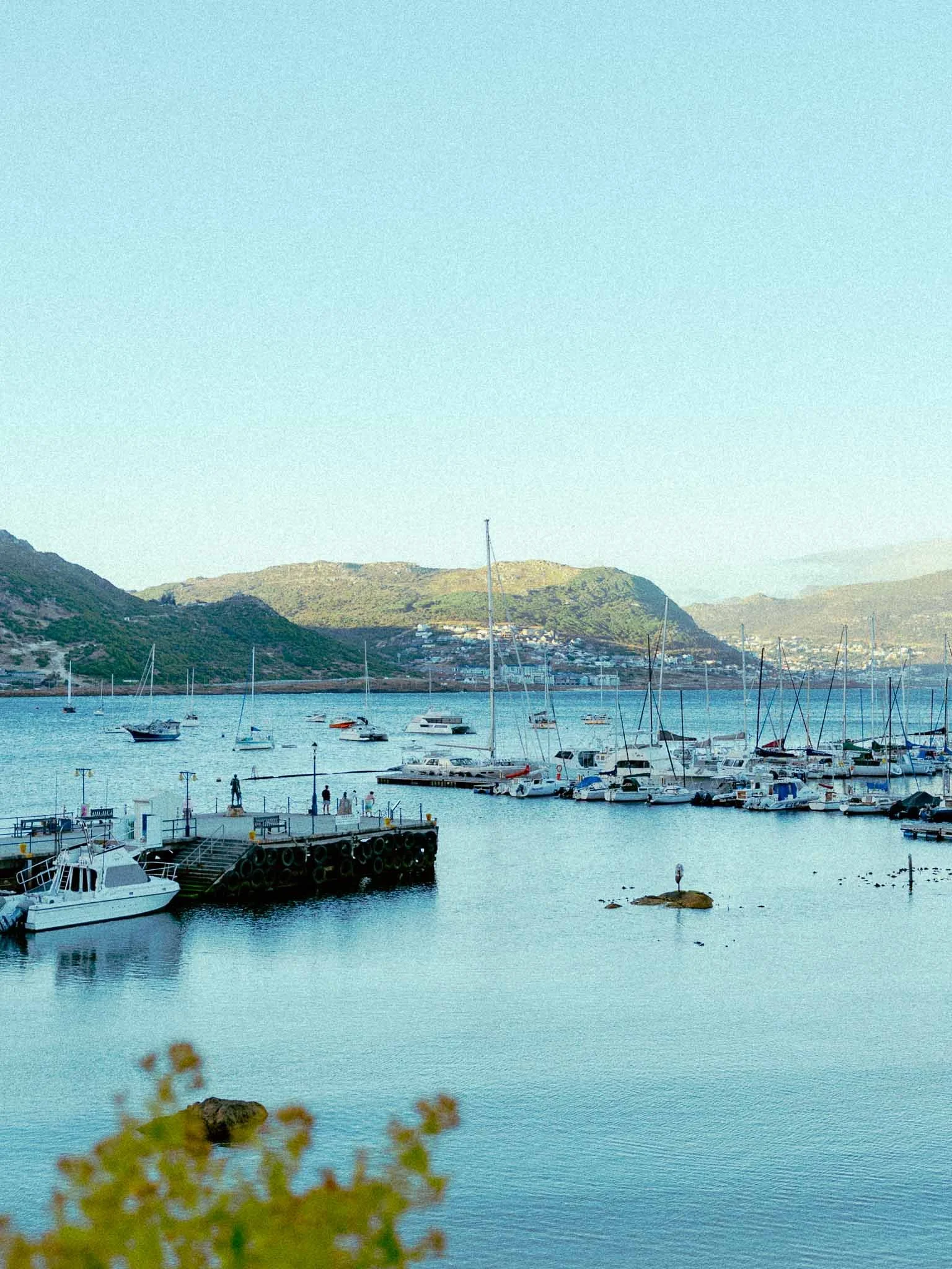 A harbor with sailboats and yachts docked, with hills in the background and a few people walking on the pier.