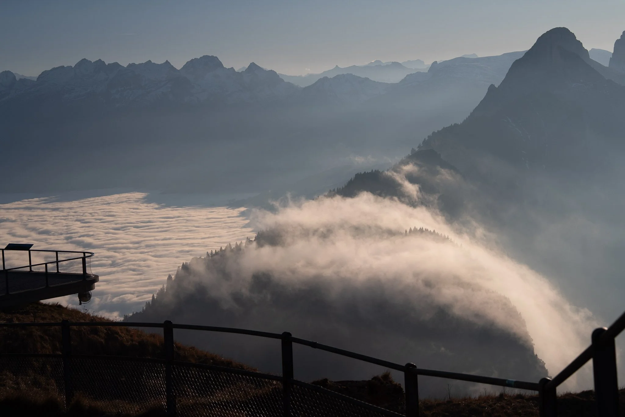Mountain landscape with clouds and mist over peaks, viewing platform and fences in foreground.