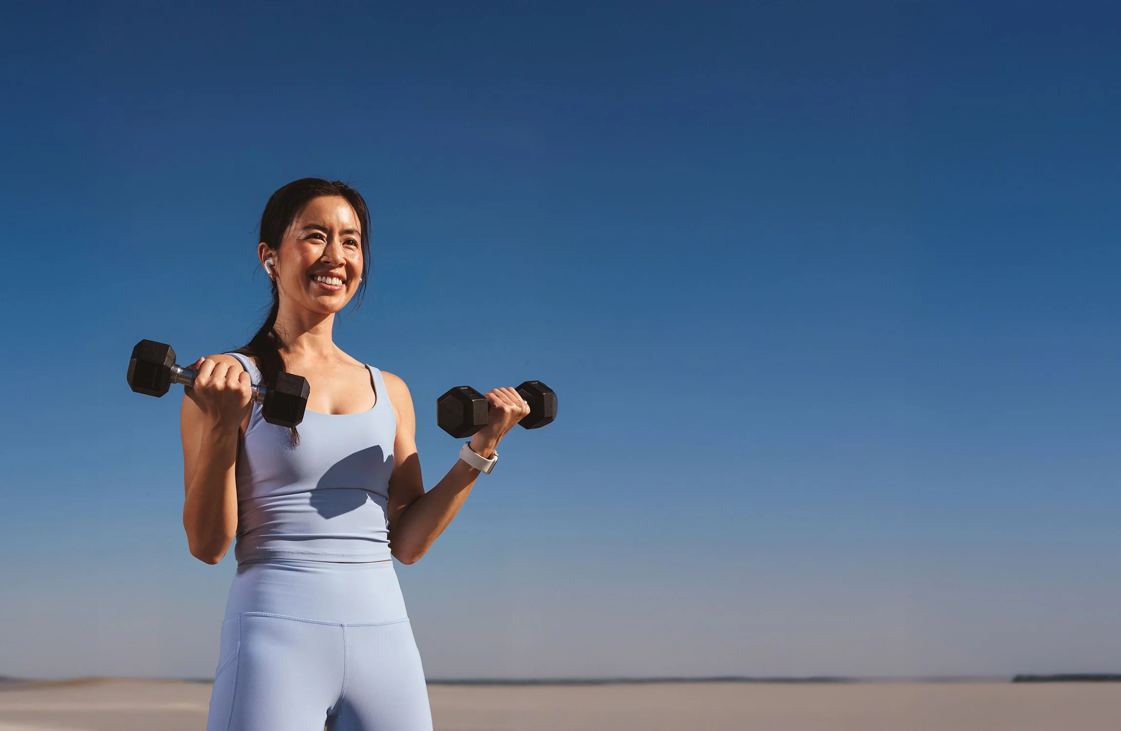 Young woman in workout attire holding dumbbells outdoors on a clear sunny day, smiling at the camera.