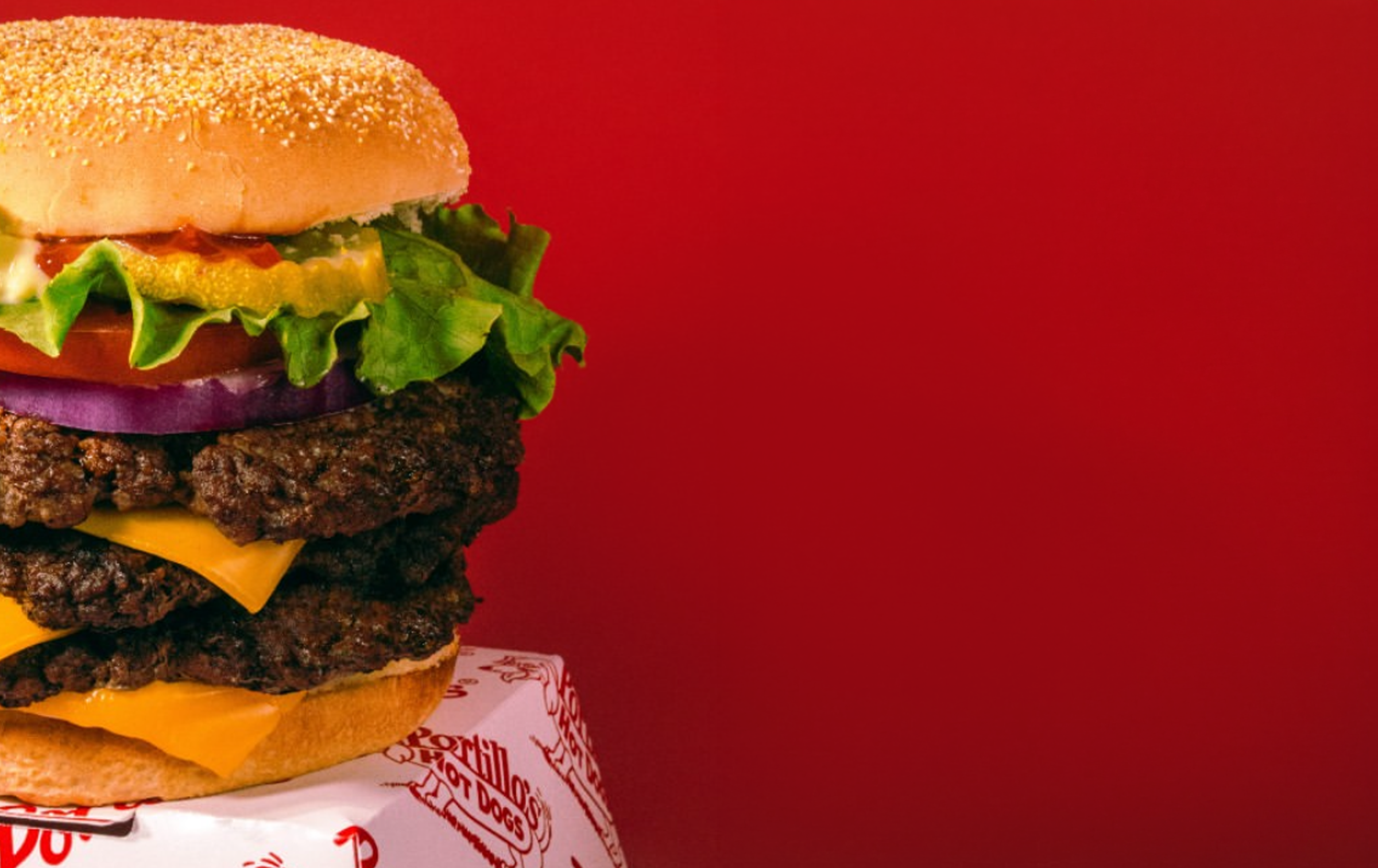 A large, multi-patty cheeseburger with lettuce, tomato, pickles, onions, cheese, and a hamburger patty between a sesame seed bun, placed on a white wrapper against a red background.