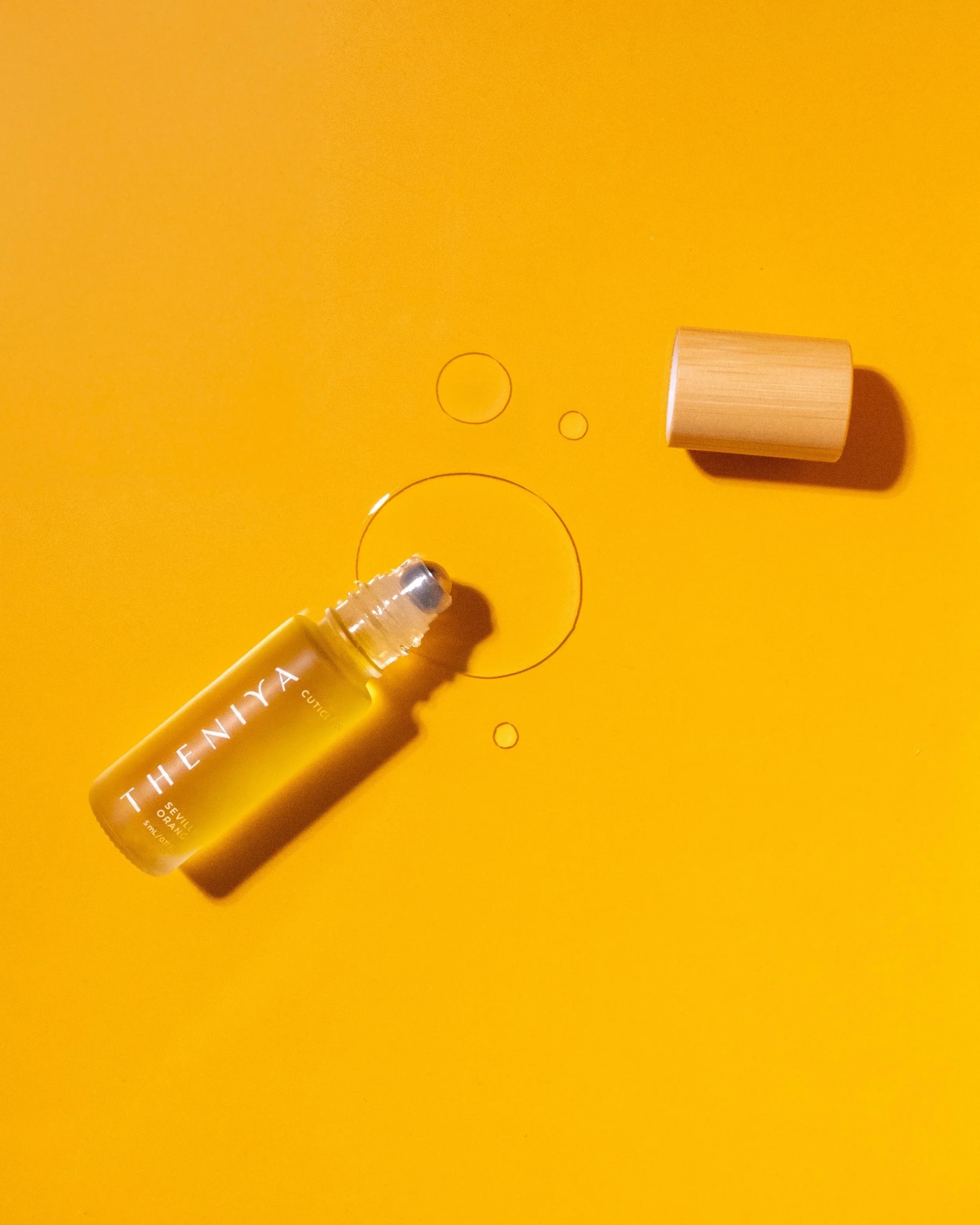 A yellow bottle of Theniya cuticle oil with silver rollerball applicator, a wooden cap, and oil on a yellow background.