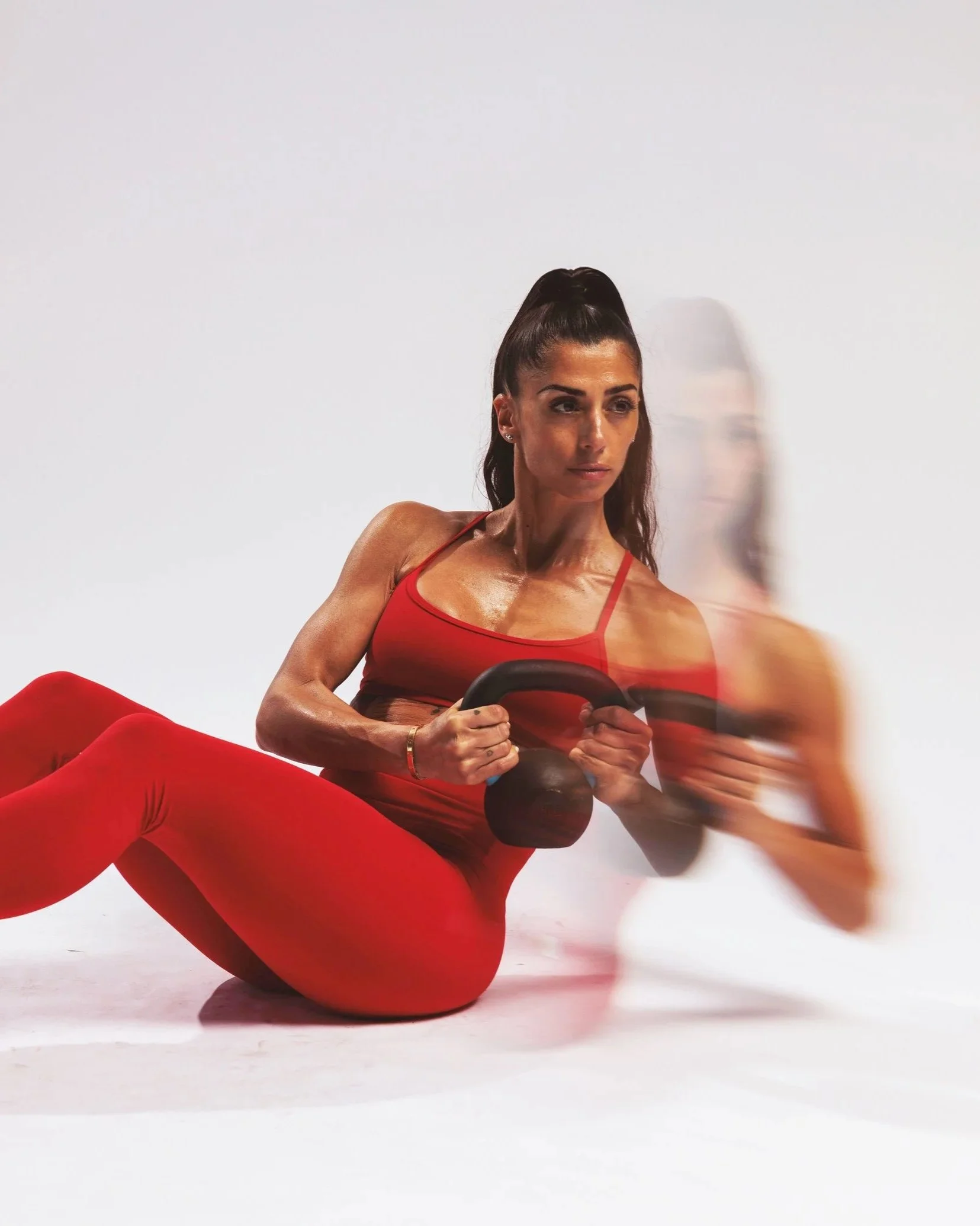 Woman in red workout clothes doing Russian twist exercise with a kettlebell, with a blurred reflection on the right side.