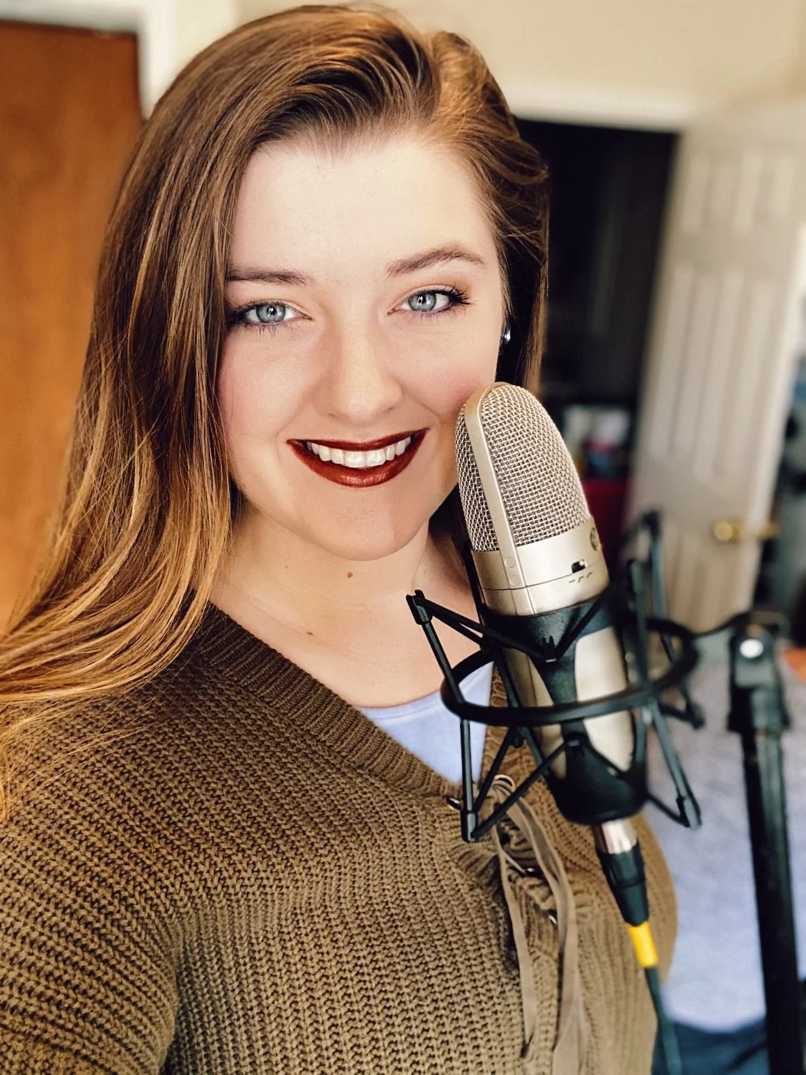 A woman with long red hair and blue eyes smiling in front of a microphone in a recording studio or home studio setting.