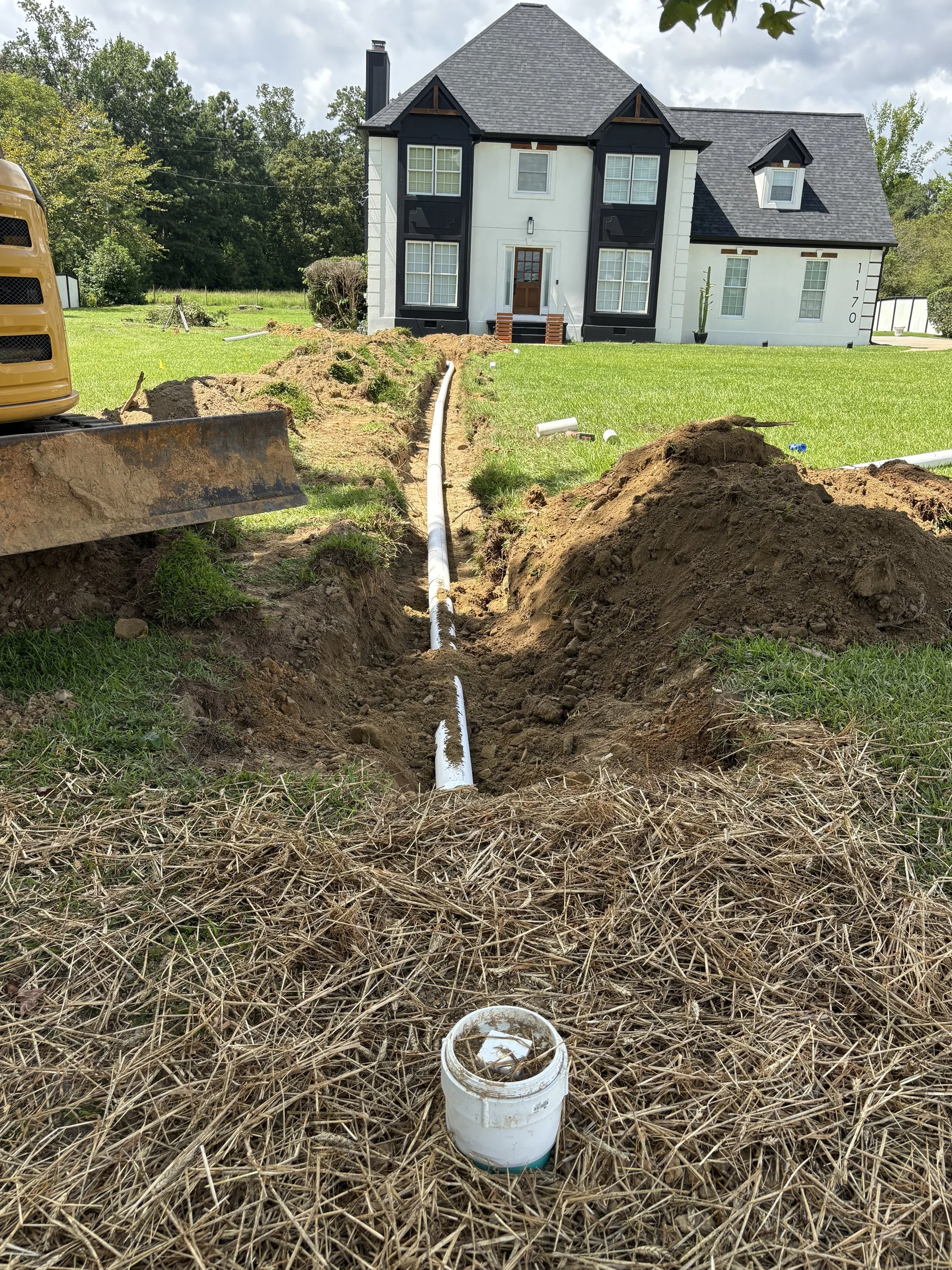 A newly installed white PVC sewer pipe laid in a freshly dug trench across a residential front lawn, showcasing a complete sewer line replacement project outside a large white house.