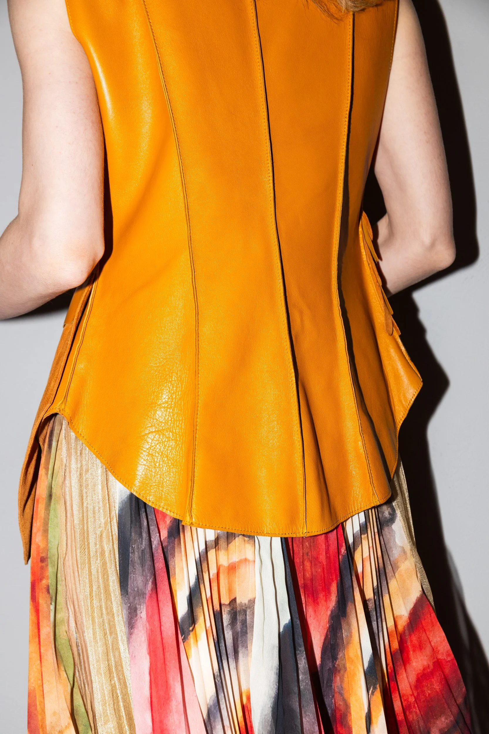 A person wearing a sleeveless, orange leather vest over a multicolored, abstract-patterned skirt.
