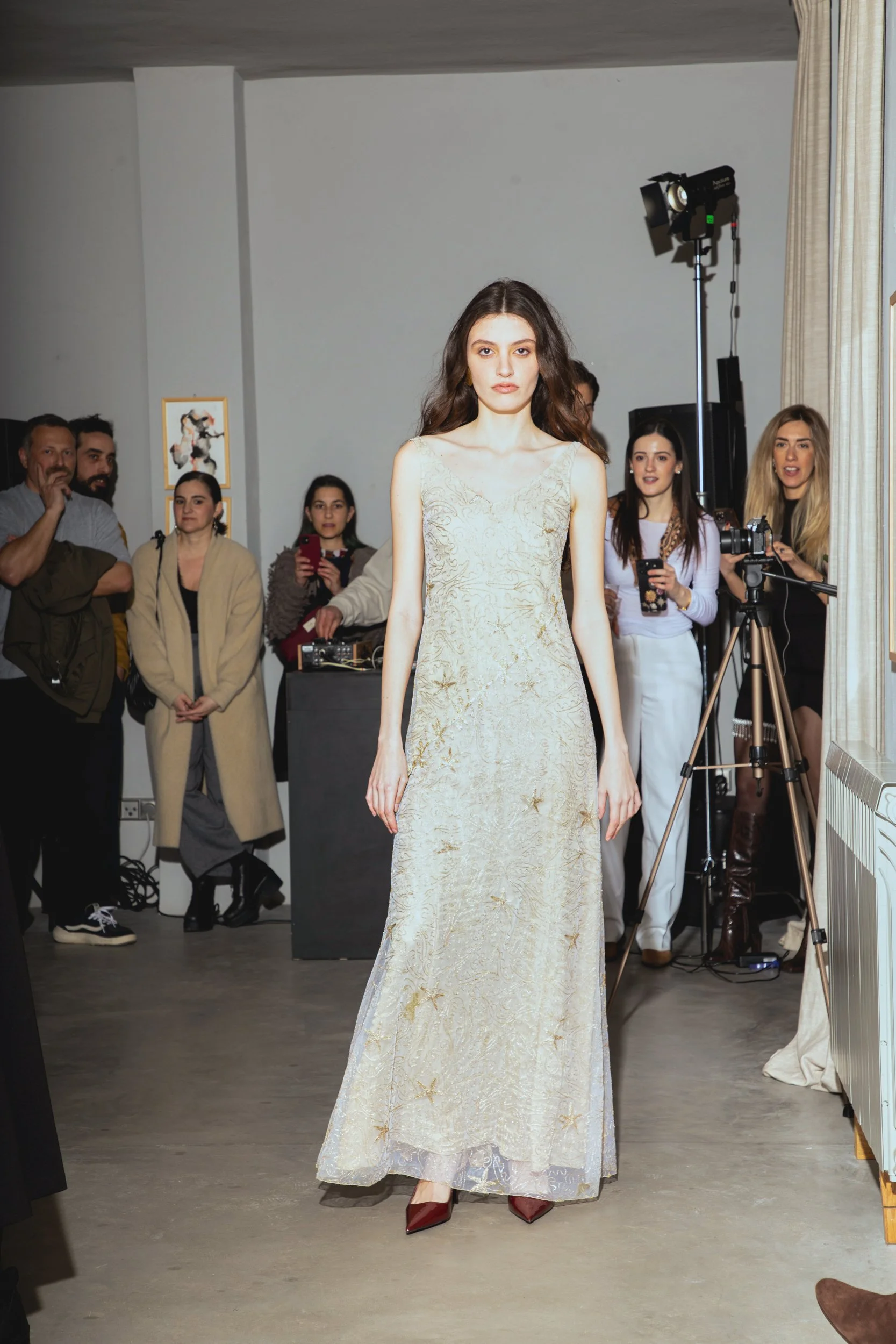 A young woman in a long, beige, embroidered dress stands on a runway at a fashion show, with an audience and photographers watching.