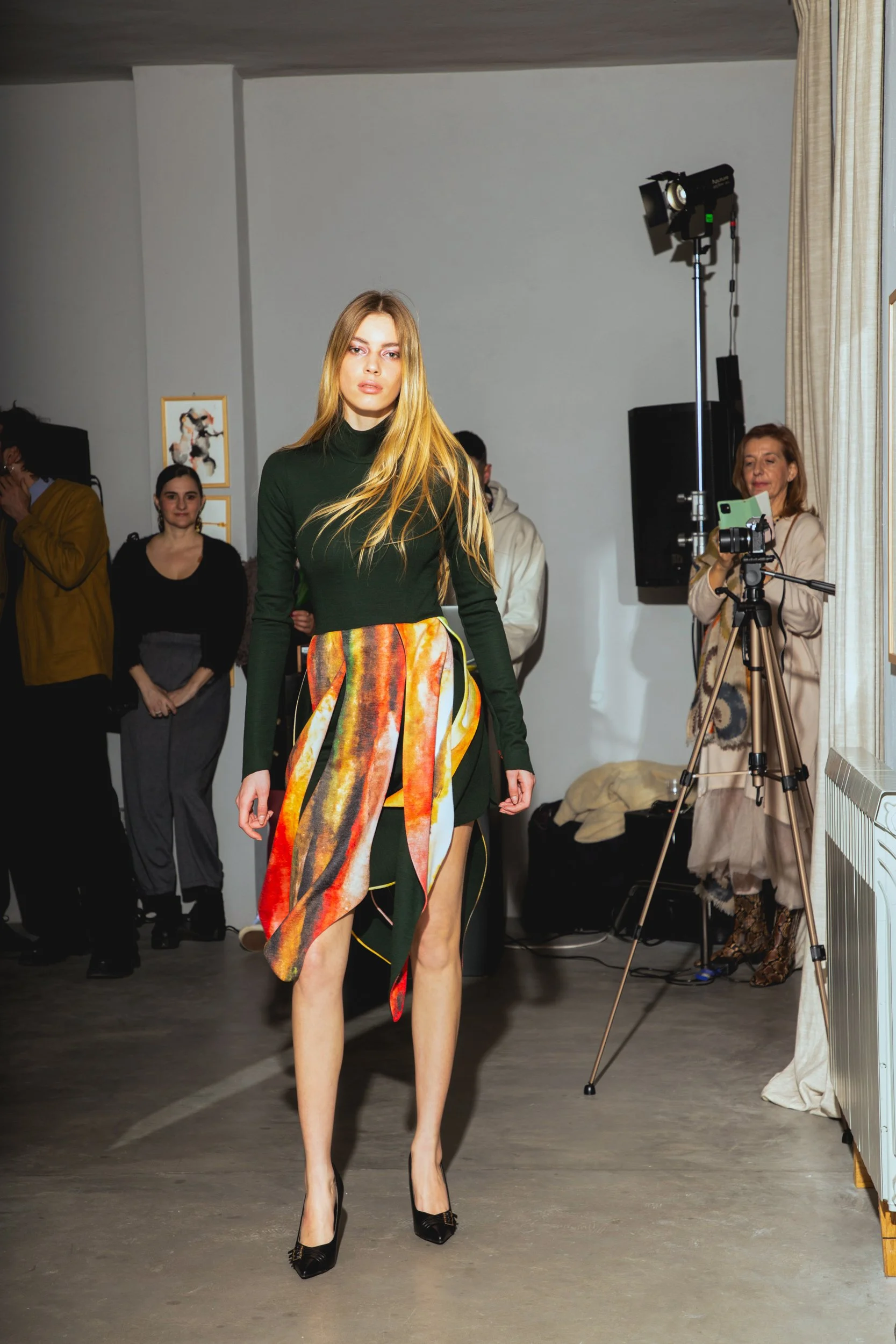 A woman walking on a runway during a fashion show, wearing a green long-sleeve dress with a colorful, patterned scarf tied around her waist and black high heels. Several people are in the background, including a photographer taking pictures.
