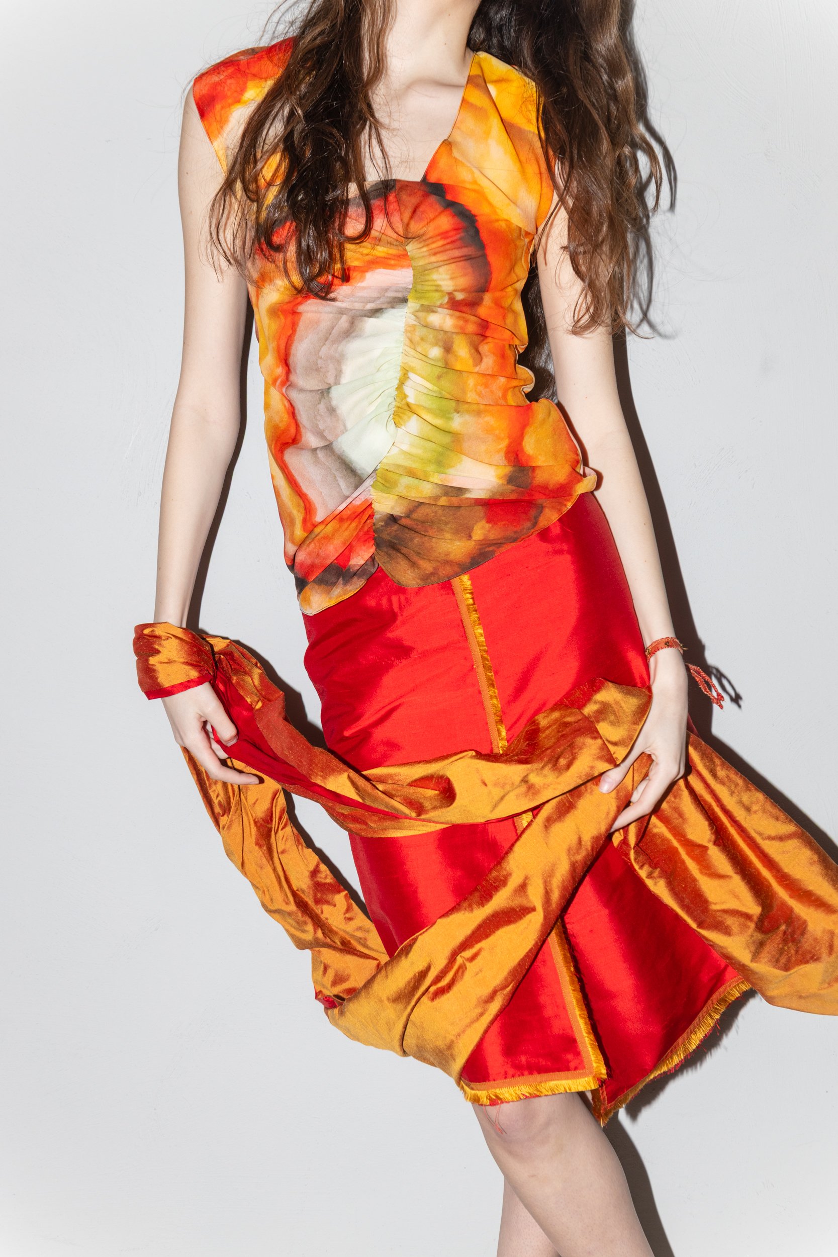 A woman wearing a colorful, artistic, abstract-patterned sleeveless top with shades of orange, red, yellow, and brown, and a matching red skirt with orange accents, holding up part of the skirt.