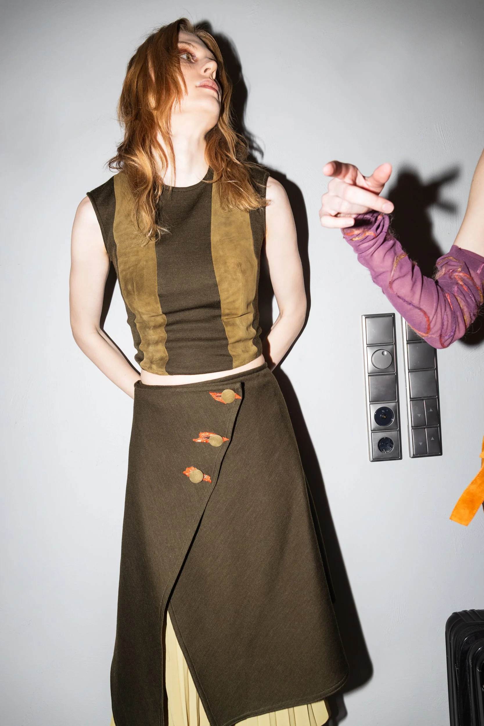 Young woman with red hair, wearing a brown and olive sleeveless top and a long dark skirt with embroidered buttons, standing against a white wall with a shadow cast to the side.